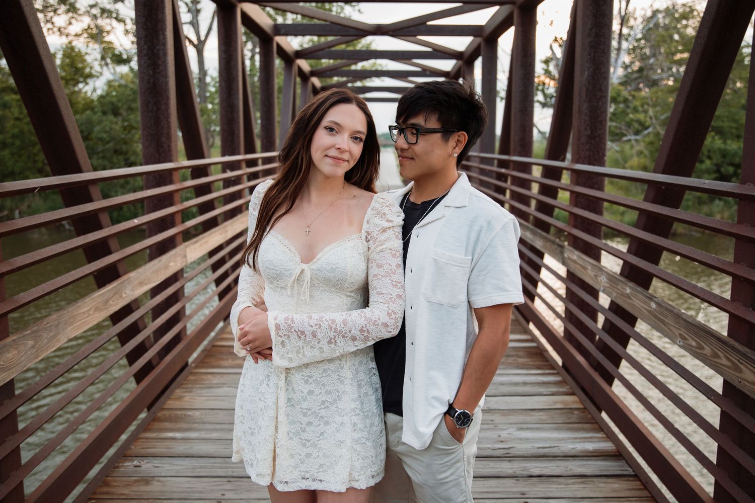 Engaged couple posing together on a wooden bridge, woman in white lace dress, man in white shirt.