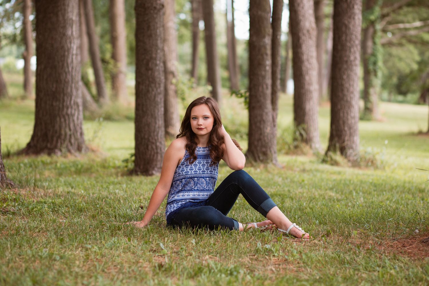 Young woman in blue top and jeans sitting on grass among tall pine trees in a serene outdoor setting.
