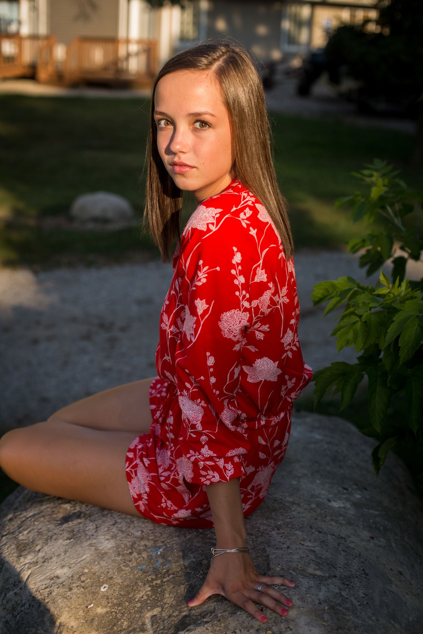 Young woman in red floral dress sitting on a rock in a garden during golden hour sunlight.