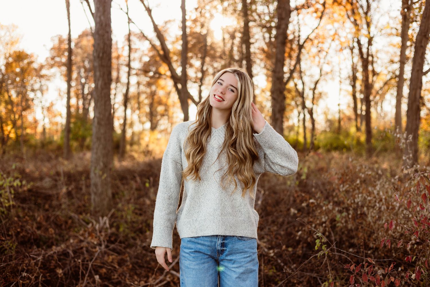Young woman in cream sweater and jeans smiling in autumn forest with golden foliage at sunset.