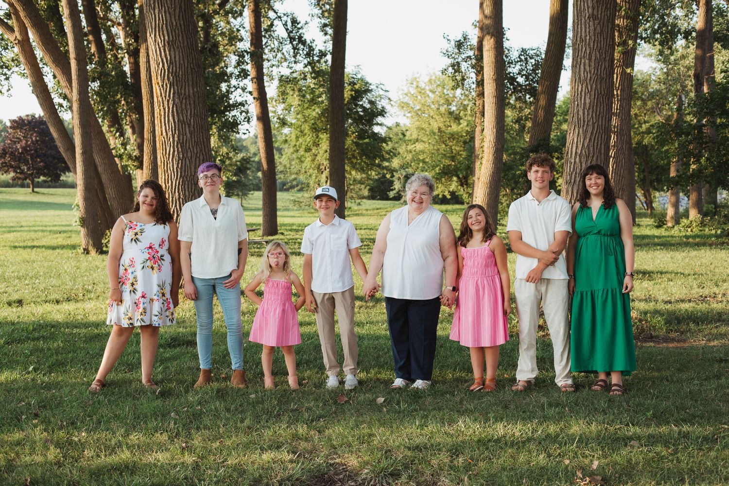 A large family group of eight people poses outdoors in a lush green park surrounded by tall trees during golden hour.