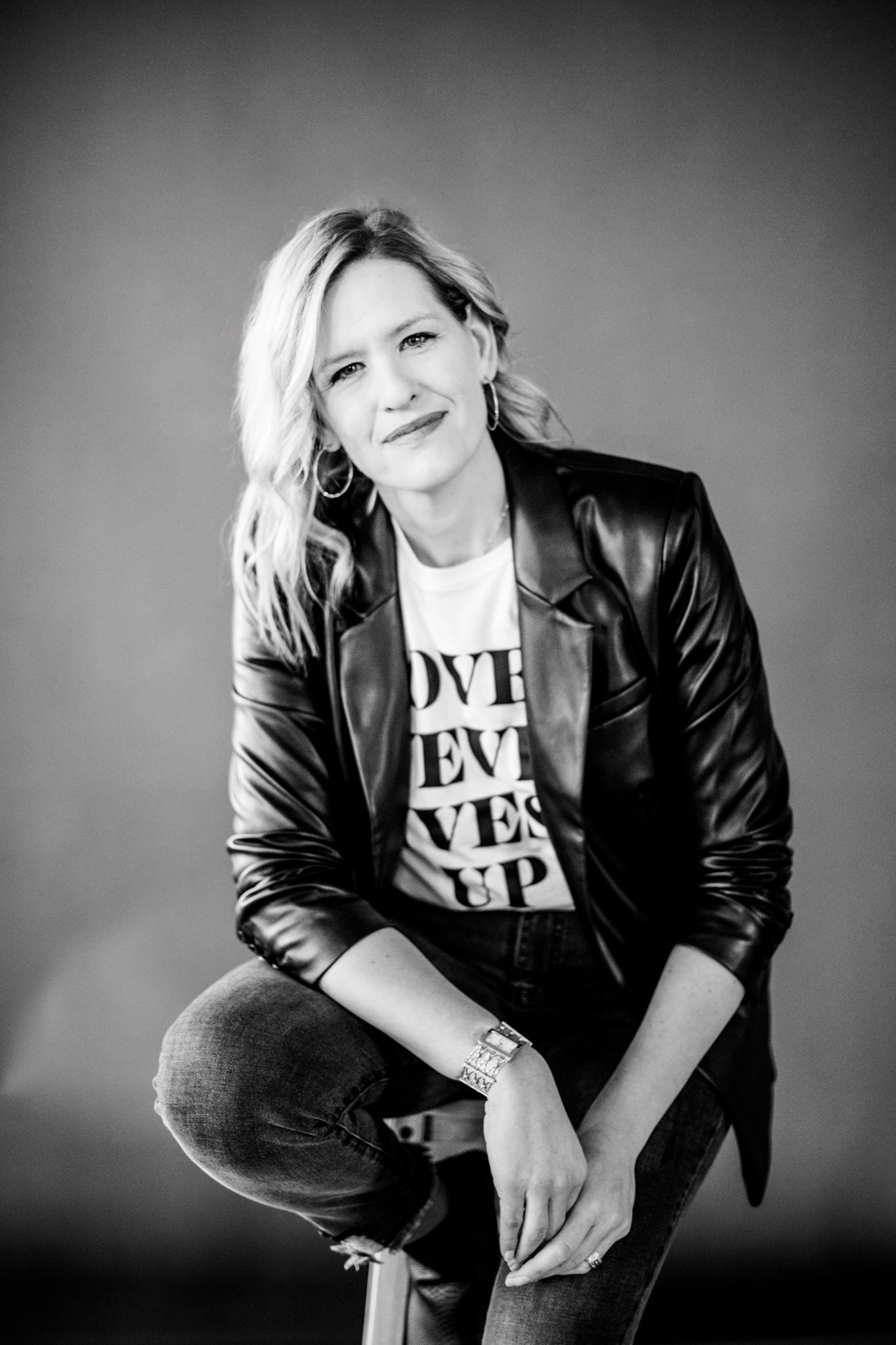 Black and white photo of smiling blonde woman in leather jacket seated on stool in studio.