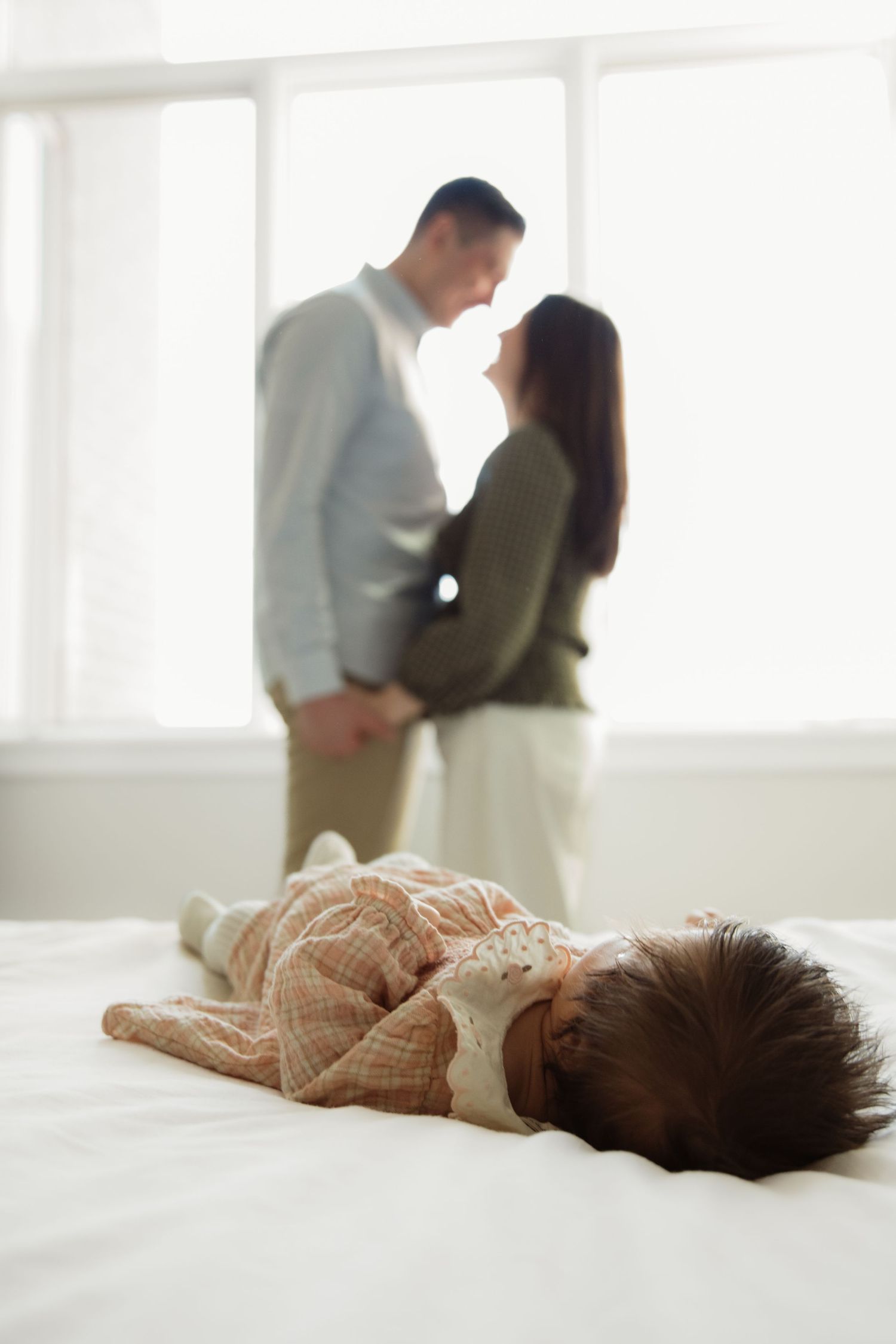 Baby lying on white bed with parents sharing romantic moment by window.