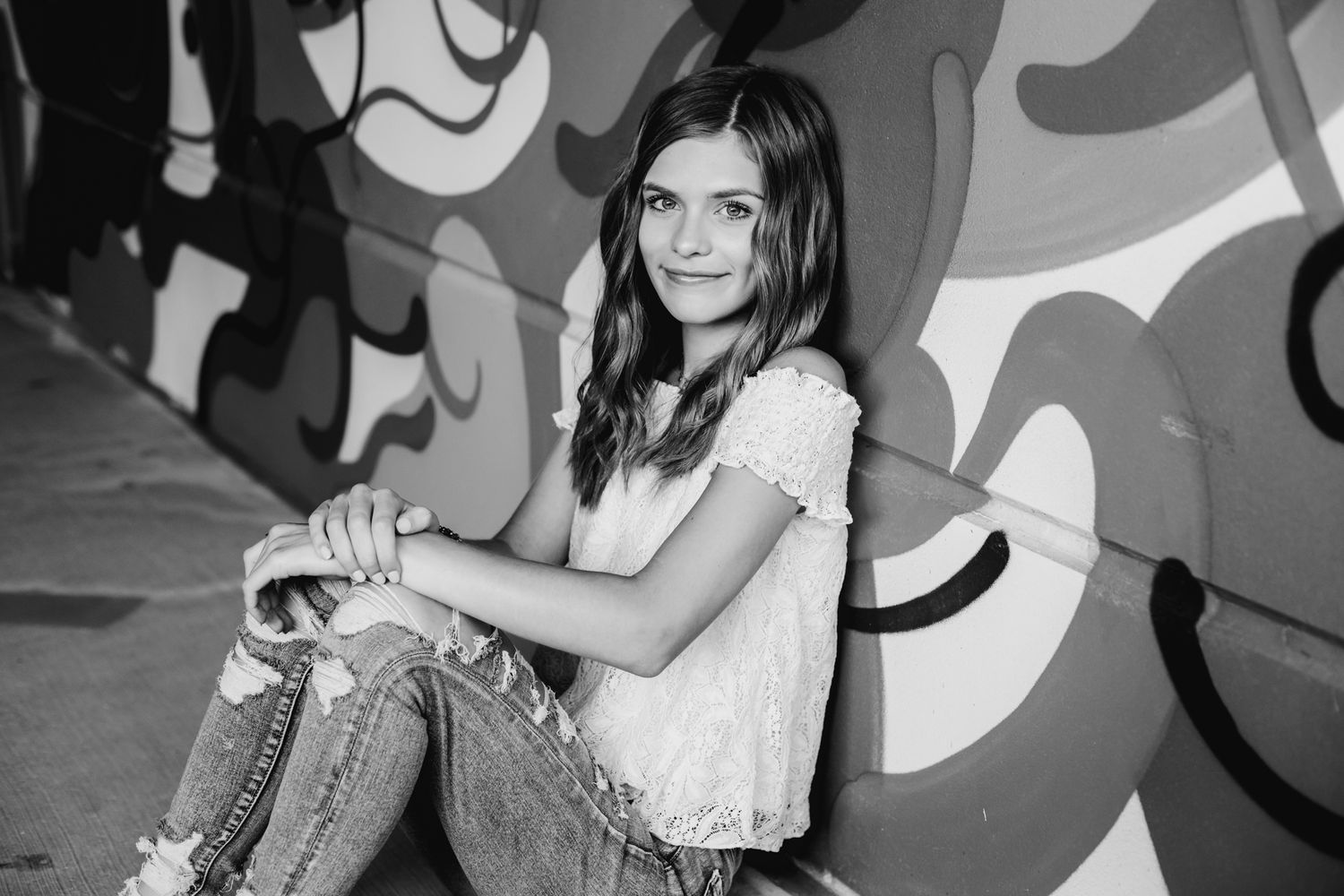 Black and white photo of young woman sitting against graffiti wall in casual outfit.