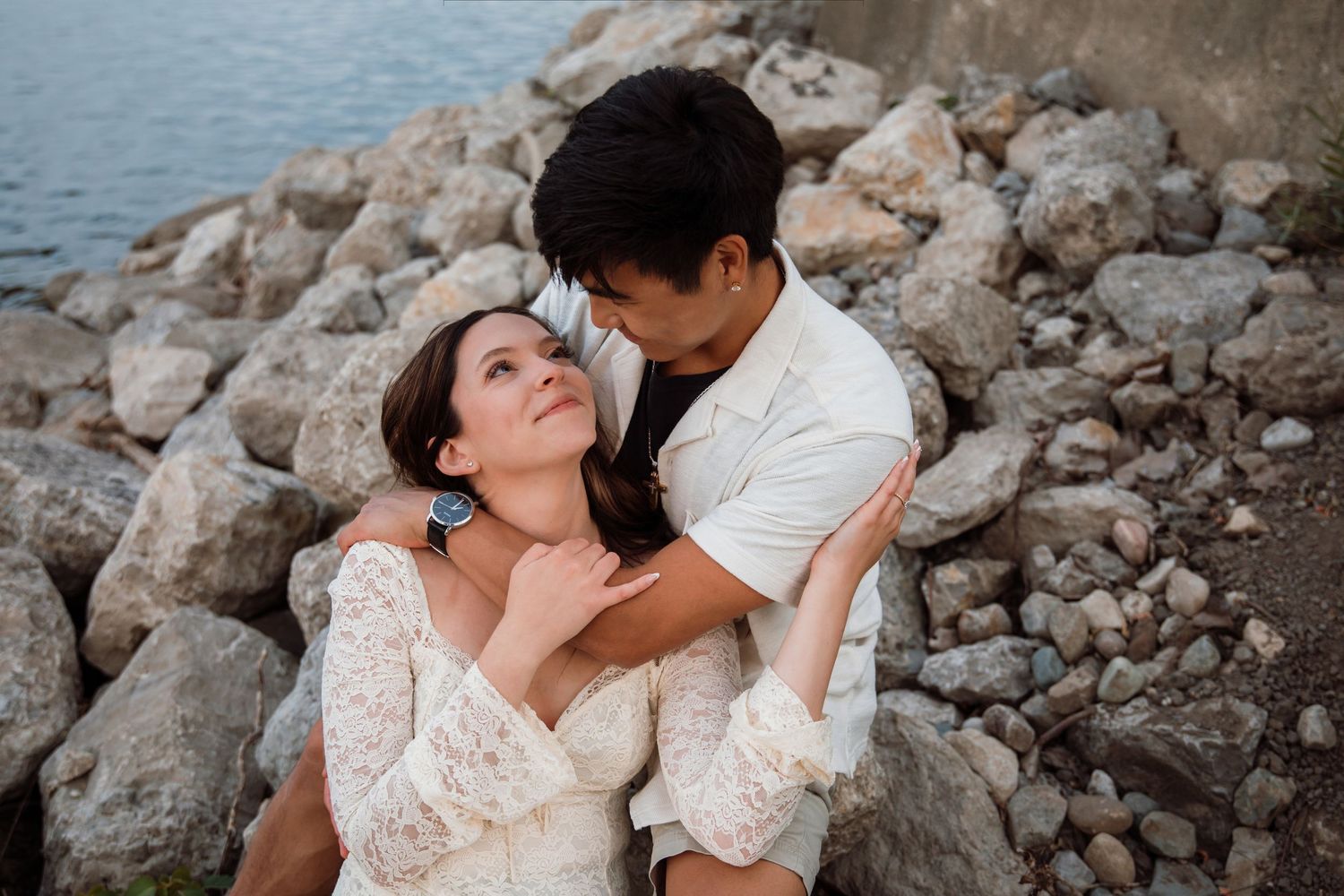 An interracial couple shares a romantic moment sitting among rocks by the water, the woman in lace gazing up at her partner.
