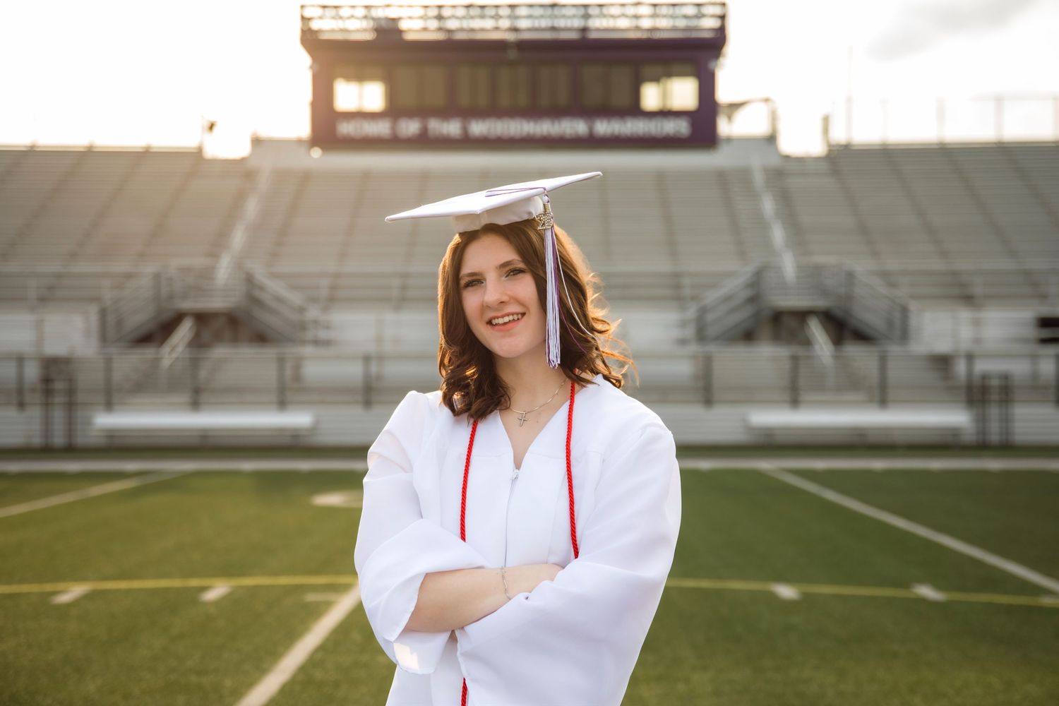 Smiling female graduate in white cap and gown stands arms crossed on football field at Woodhaven Warriors stadium.
