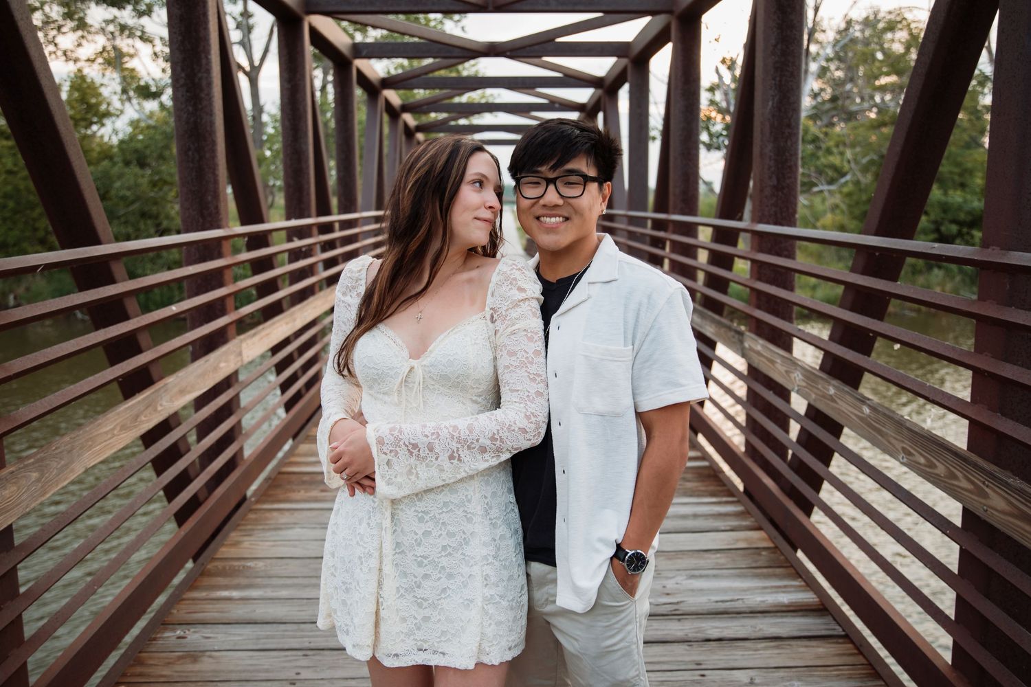 Engaged couple standing together on a rustic wooden bridge, smiling softly in natural light.