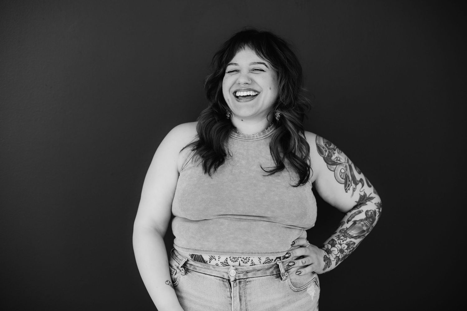 Black and white photo of a laughing woman with tattoos in a sleeveless top and jeans.