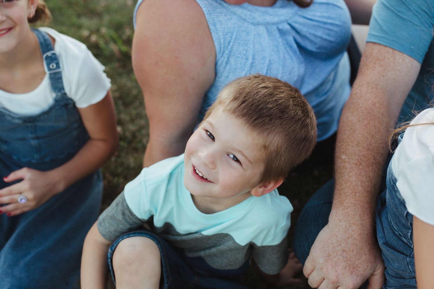 A smiling young boy in a striped teal shirt sits between family members outdoors during a casual family photo session.