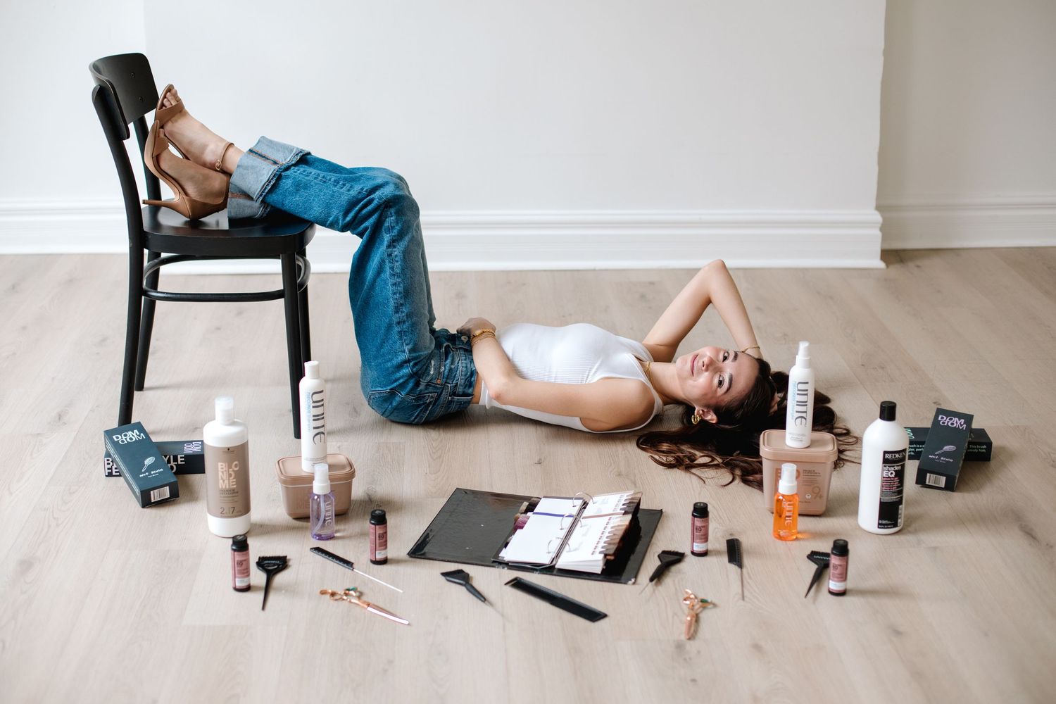 Woman lying on floor surrounded by hair care products, tools, and a black chair.