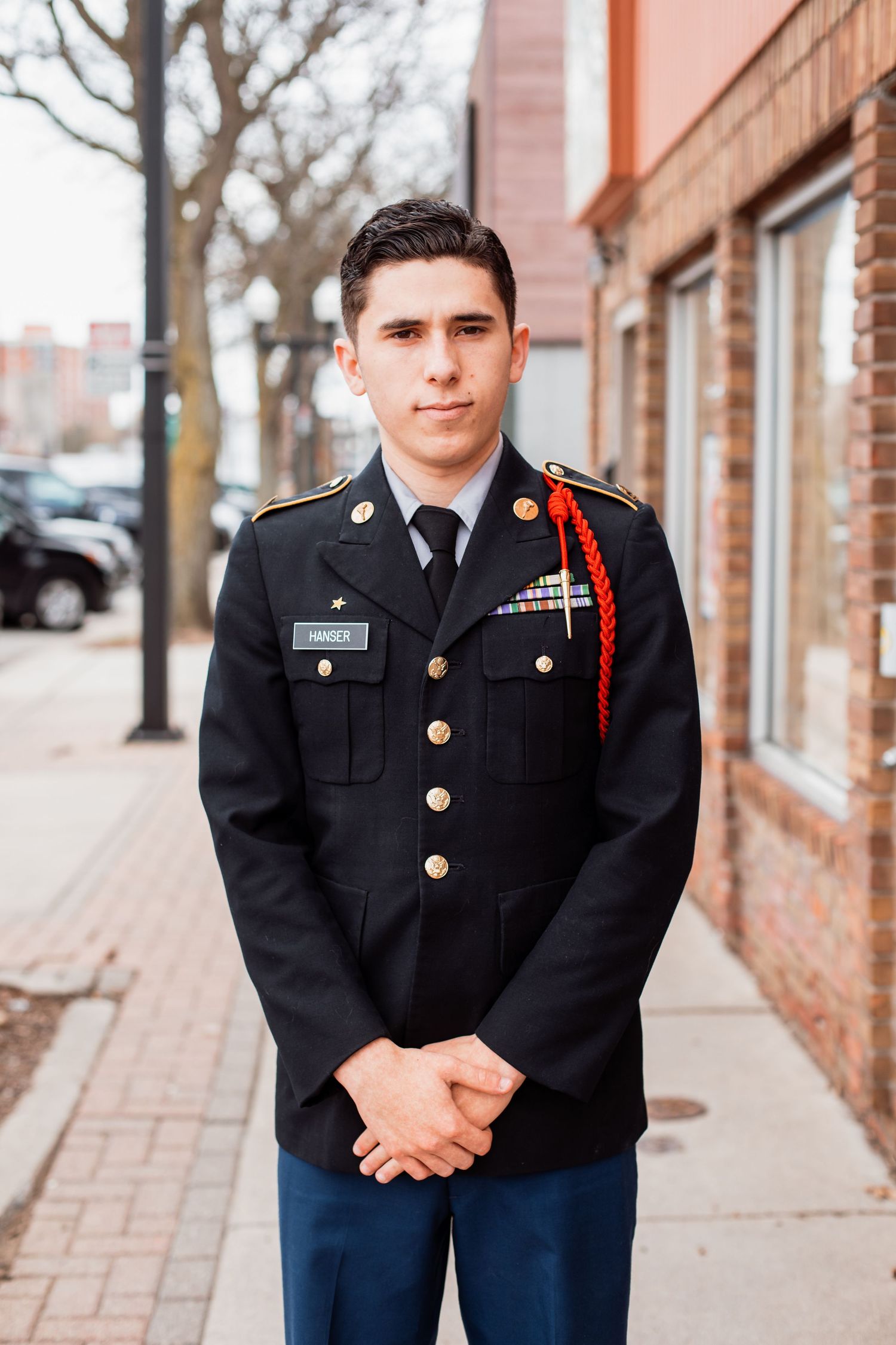 Young man in black military dress uniform with gold buttons and red cord stands on a city sidewalk.