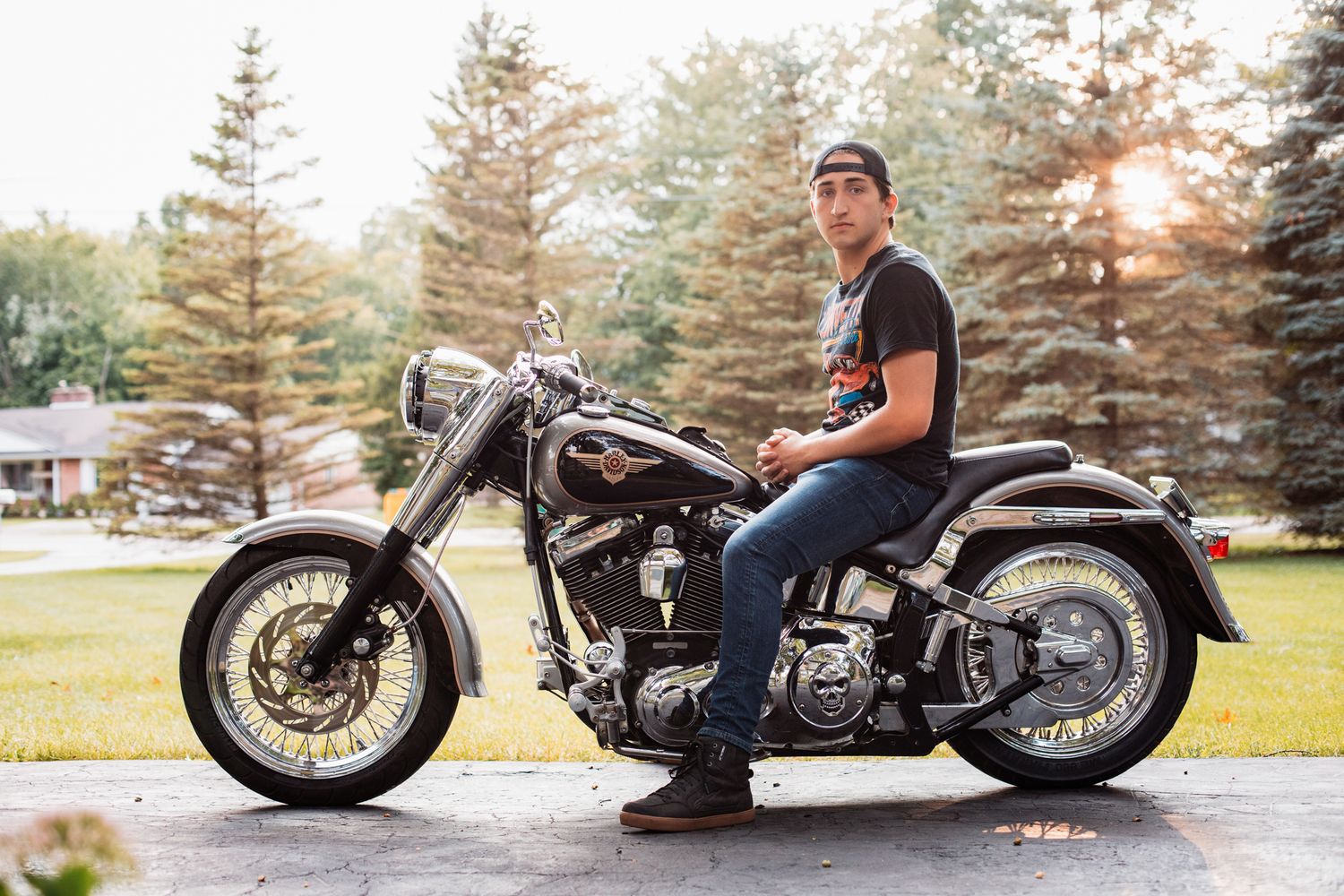 Man in black t-shirt and jeans sitting on a Harley-Davidson motorcycle in a driveway surrounded by trees.