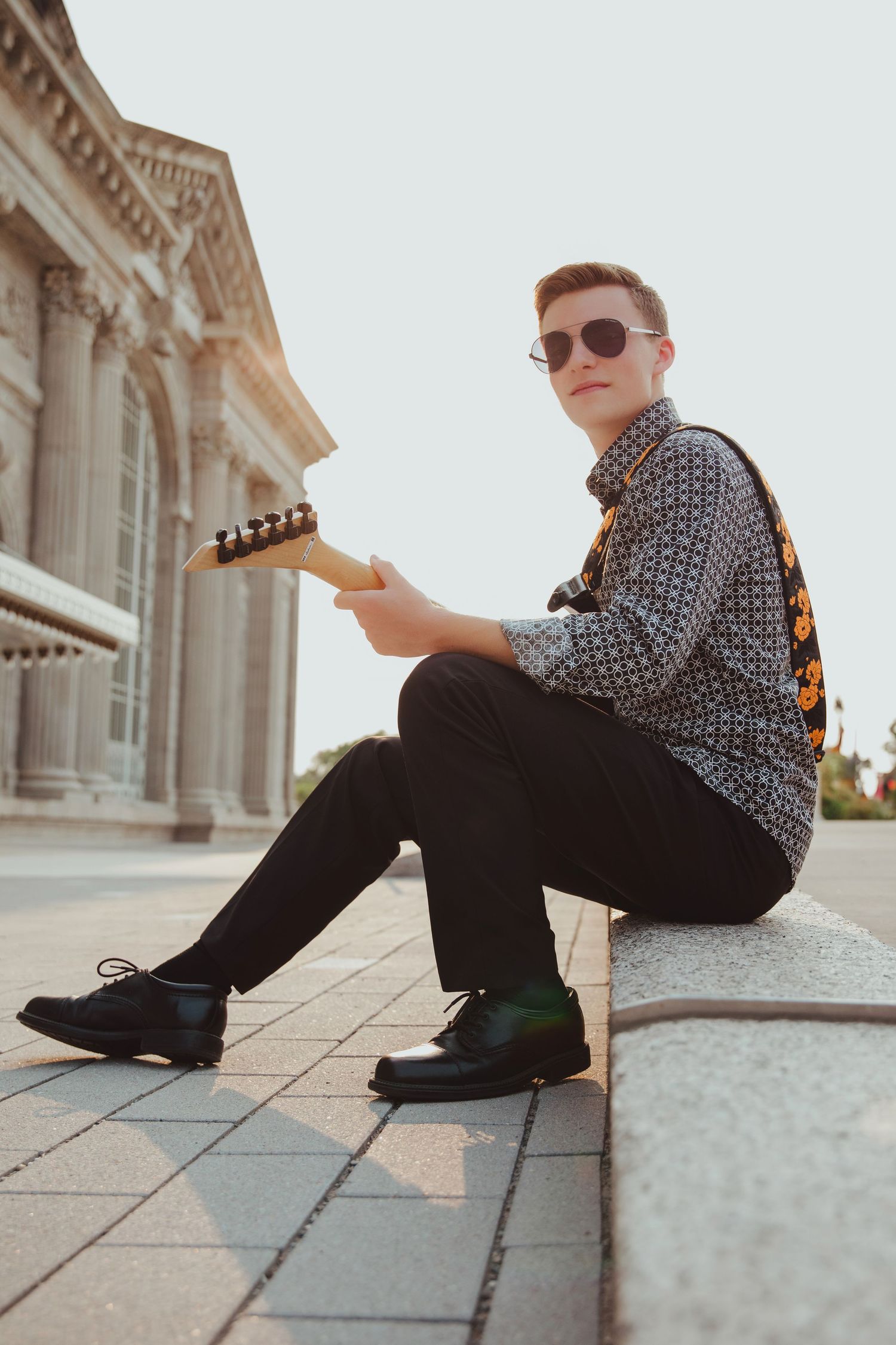 Young man in patterned shirt and sunglasses sitting on steps playing electric guitar outdoors near classical building.