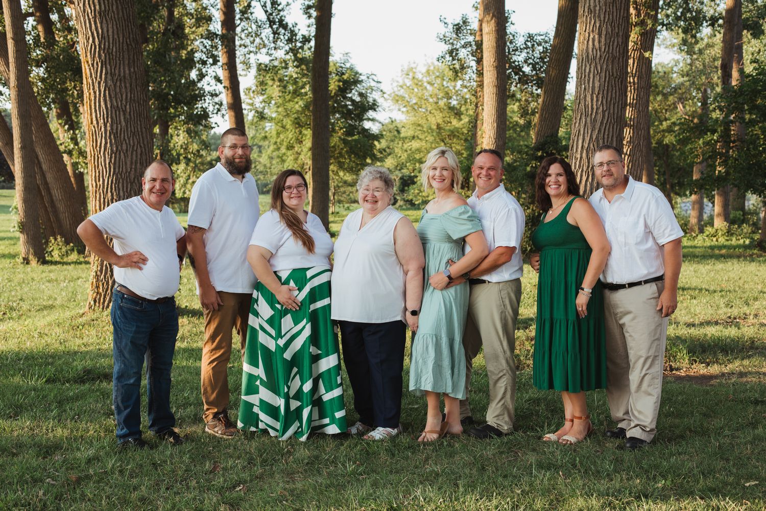A joyful family group of eight poses outdoors in a lush green park, wearing coordinated green and white outfits.