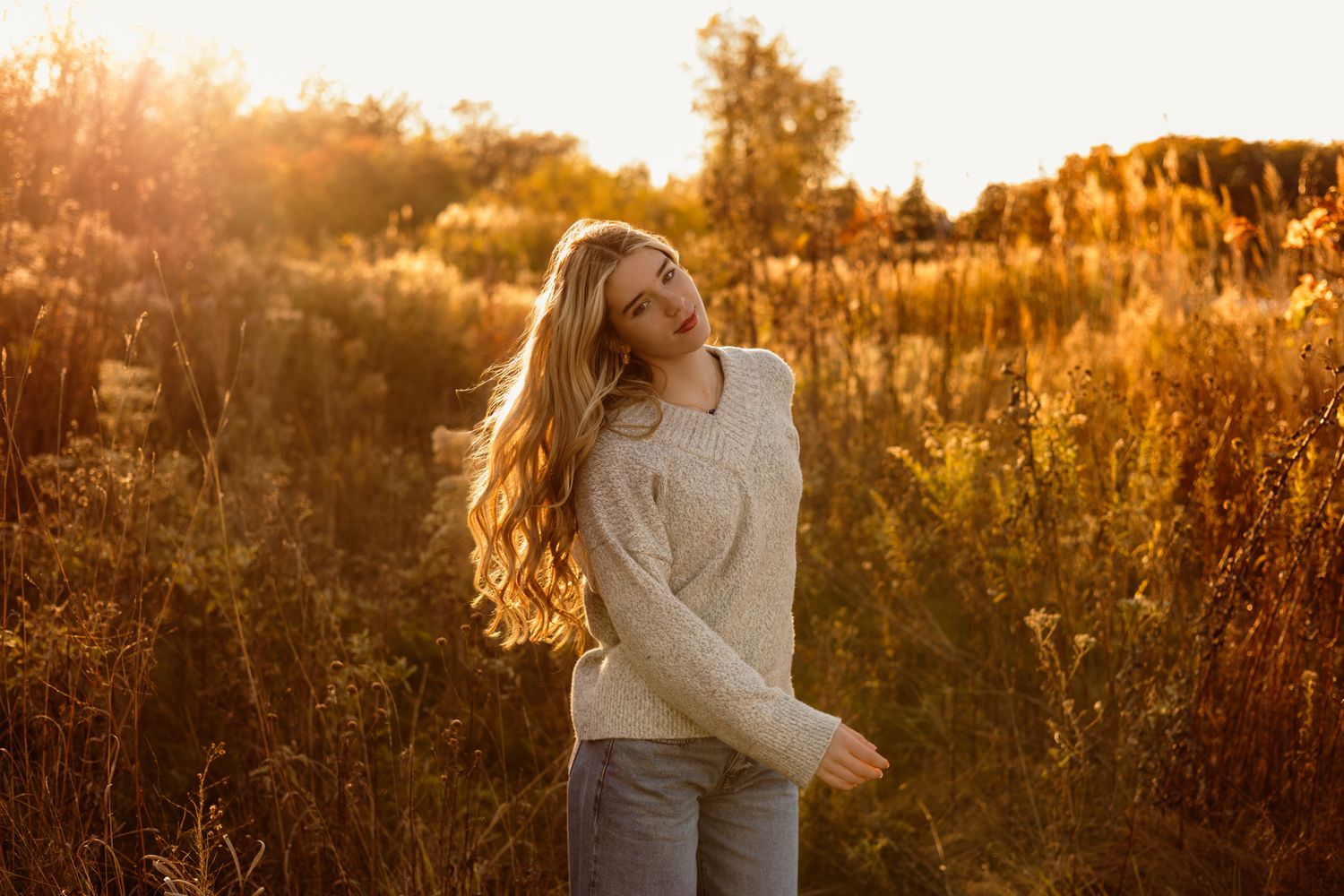 Young woman with long blonde hair in a cream sweater standing in a golden sunlit field during autumn sunset.