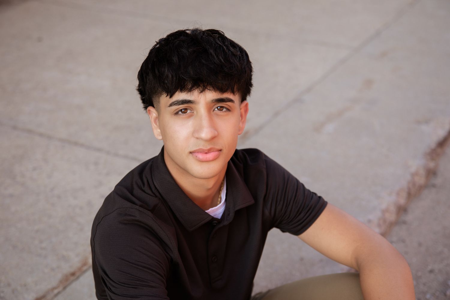 Young man with dark hair wearing a brown polo shirt sitting outdoors on a sunny day.