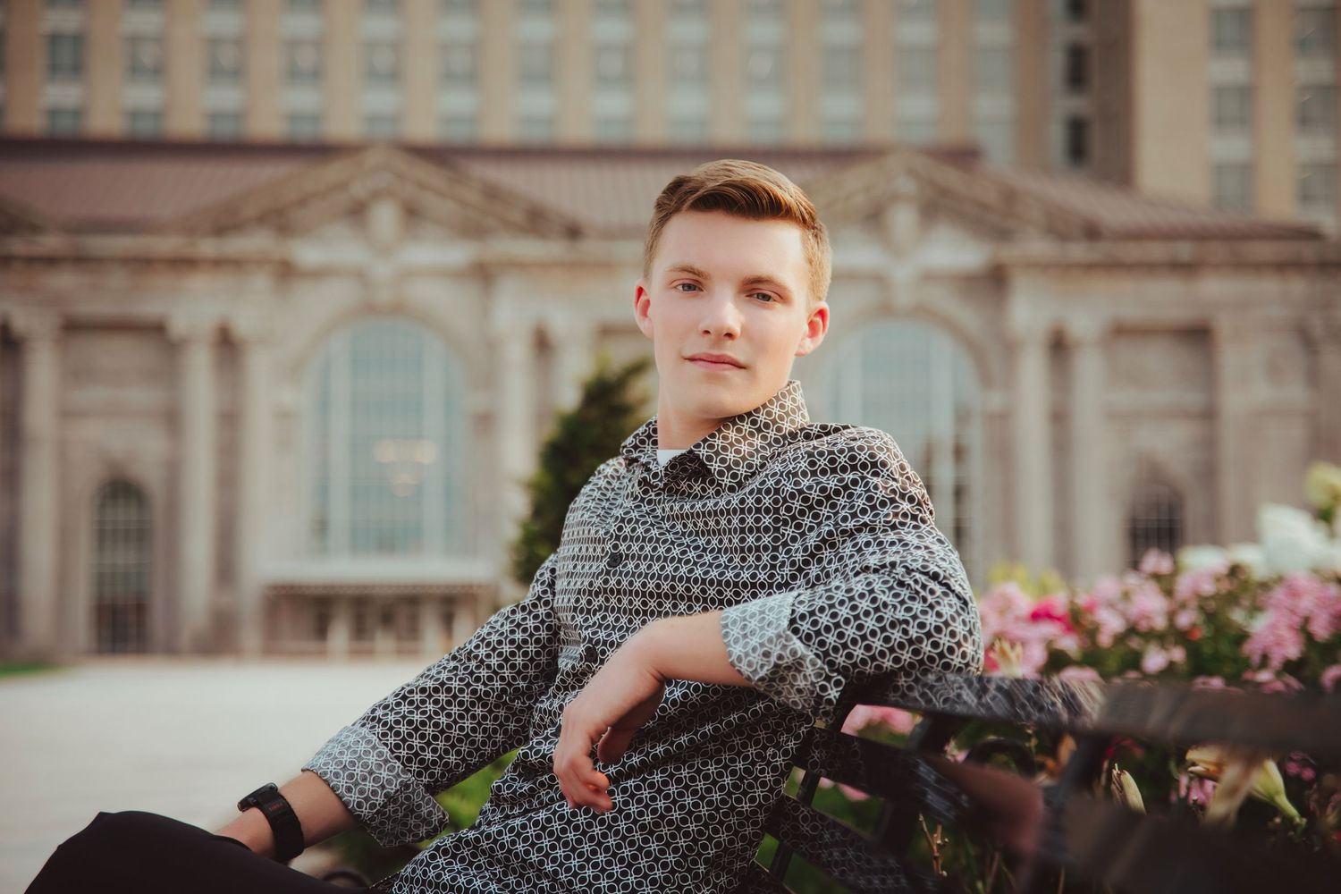 Young man in a patterned shirt sits relaxed on a bench in front of a grand building.