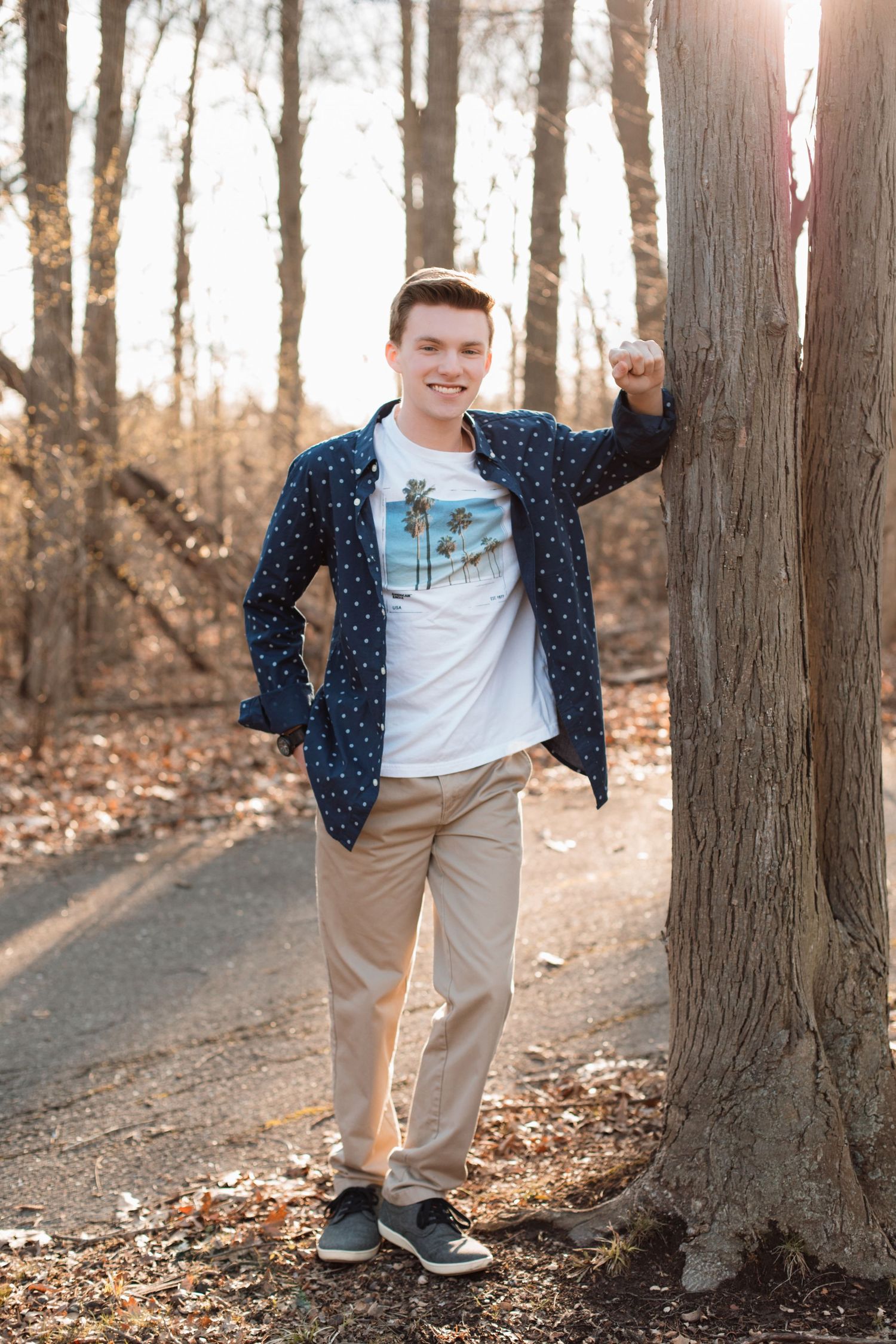 Cheerful young man in navy polka dot shirt poses beside tree on wooded path in fall setting.