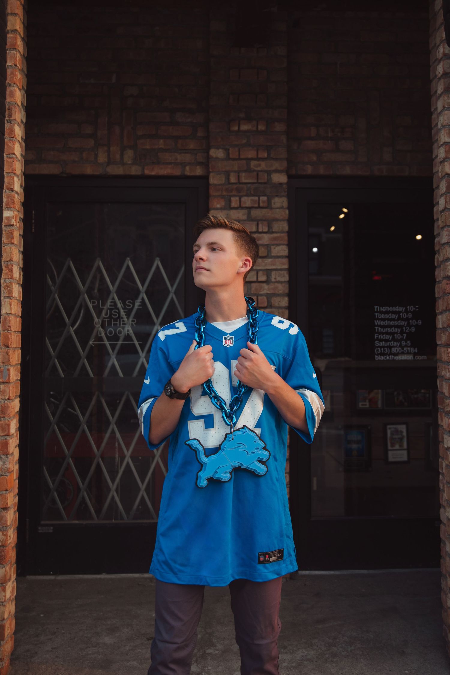 Young man in blue Lions NFL jersey posing thoughtfully in front of brick building doorway.