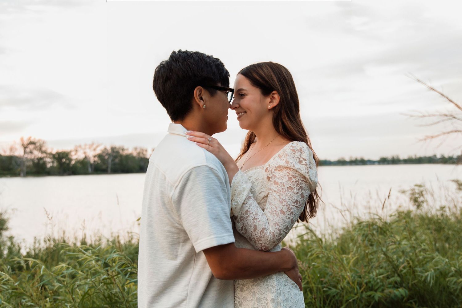 A loving interracial couple nearly kisses while embracing near a lake surrounded by tall green grass at sunset.