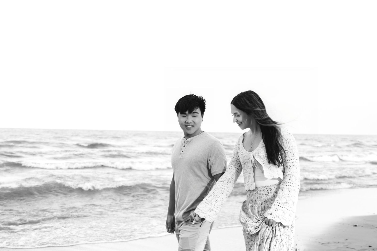 Black and white photo of a couple holding hands and smiling on a beach with ocean waves in the background.