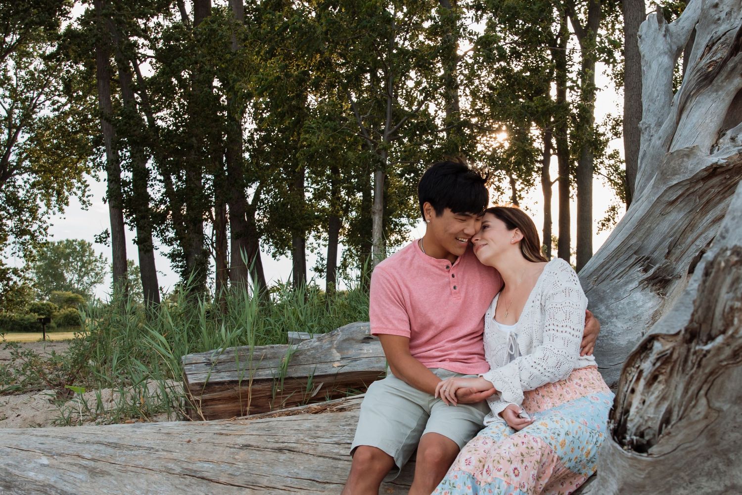 Couple sitting close together on driftwood by a beach, surrounded by lush green trees at sunset.