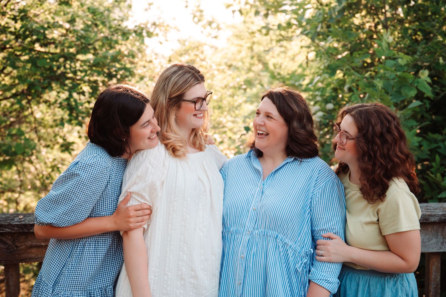 Four women laughing and embracing each other outdoors on a wooden deck with green foliage behind them.