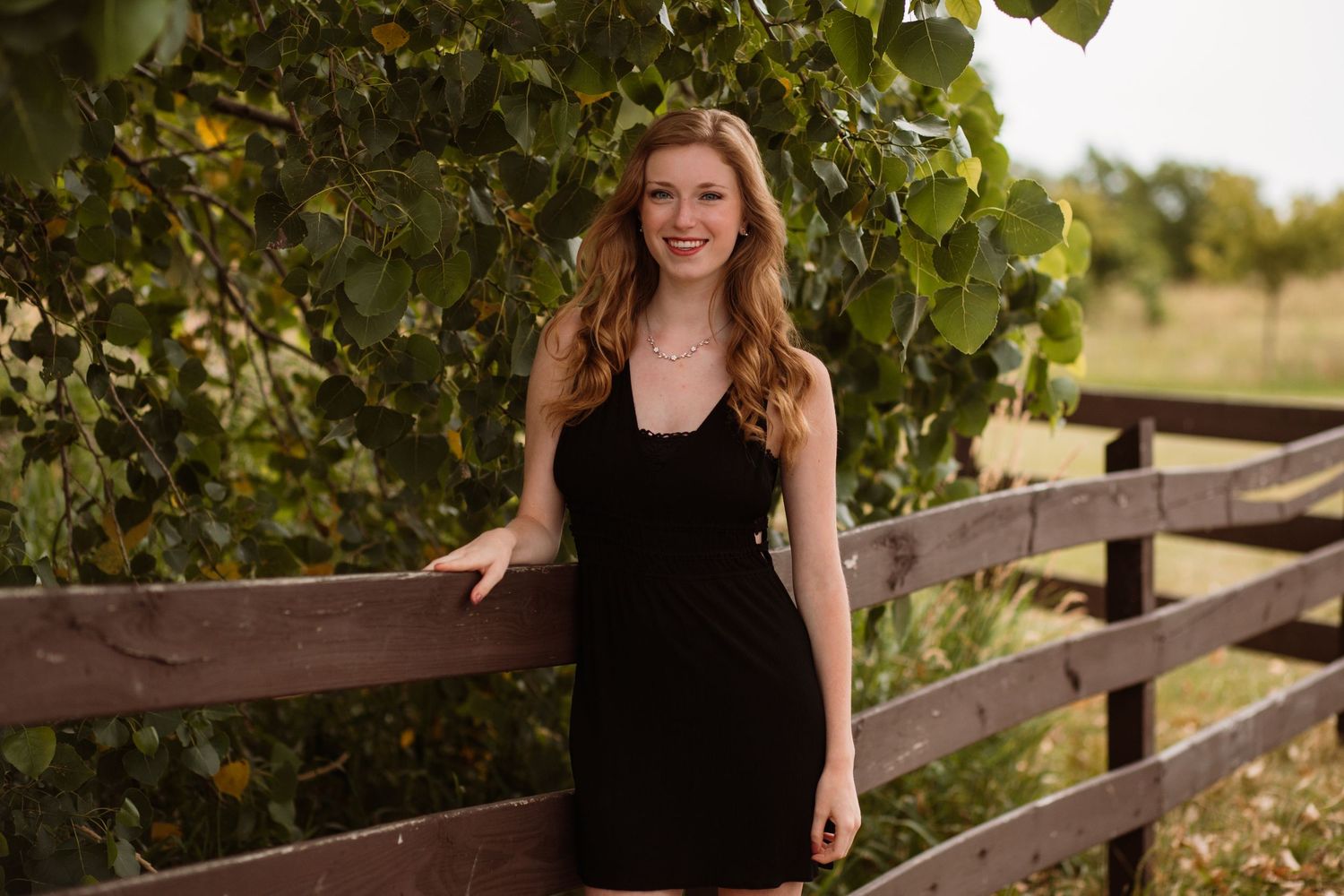 Young woman in black dress smiling by wooden fence surrounded by lush green trees outdoors.