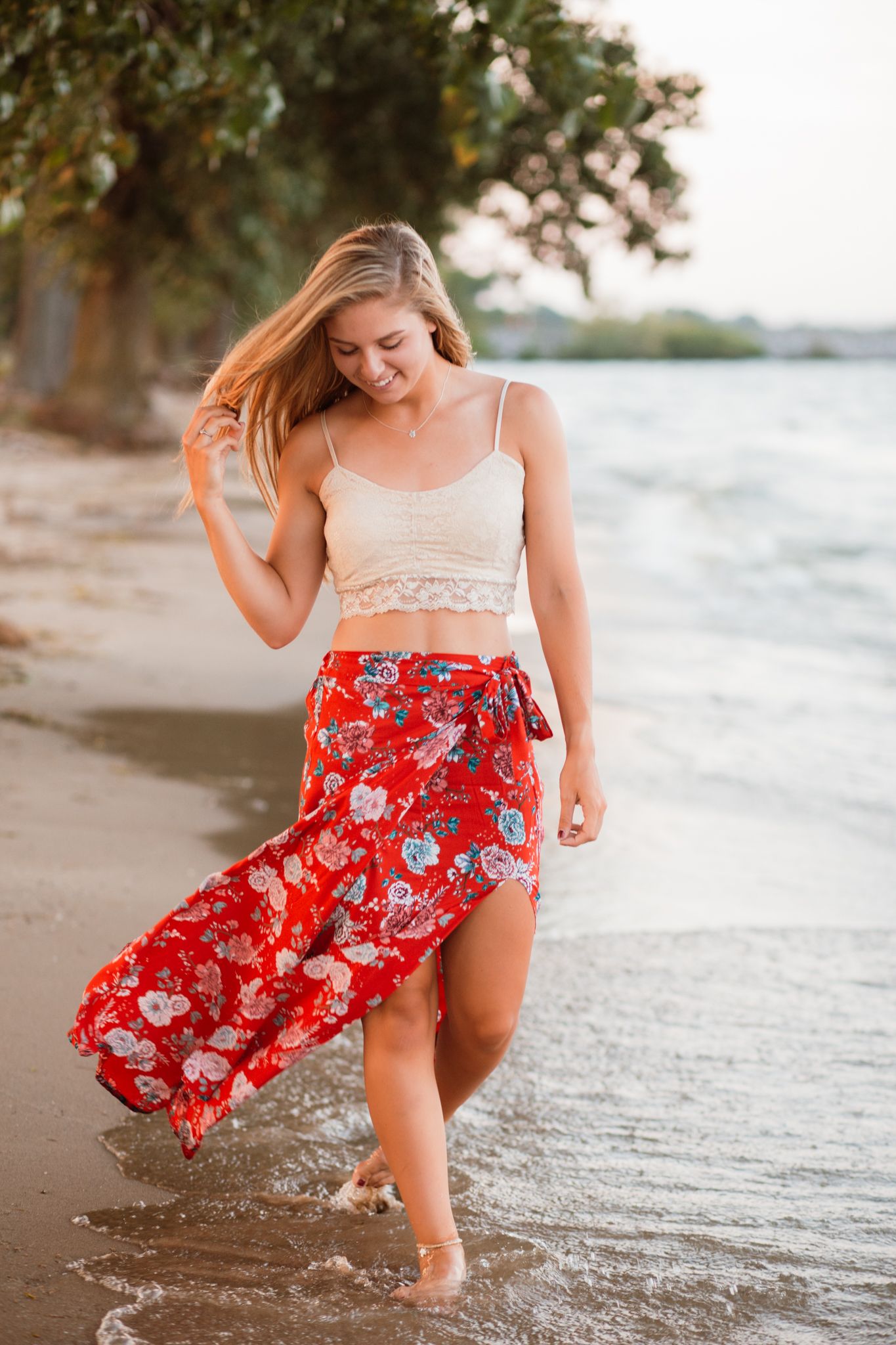 Smiling blonde woman in white crop top and red floral skirt walking along sunny beach.