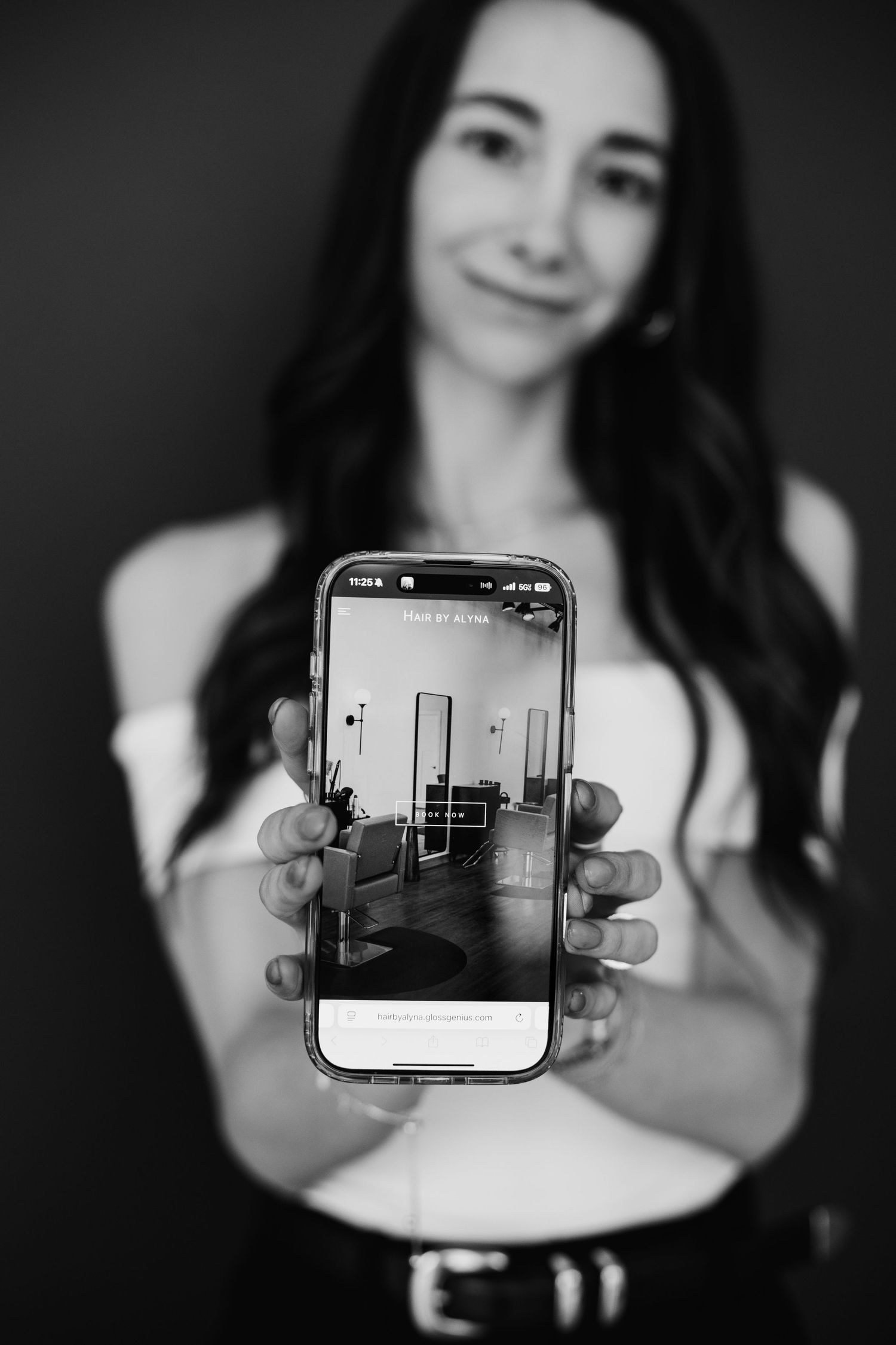 Woman in black and white photo holding smartphone displaying salon interior image toward camera.