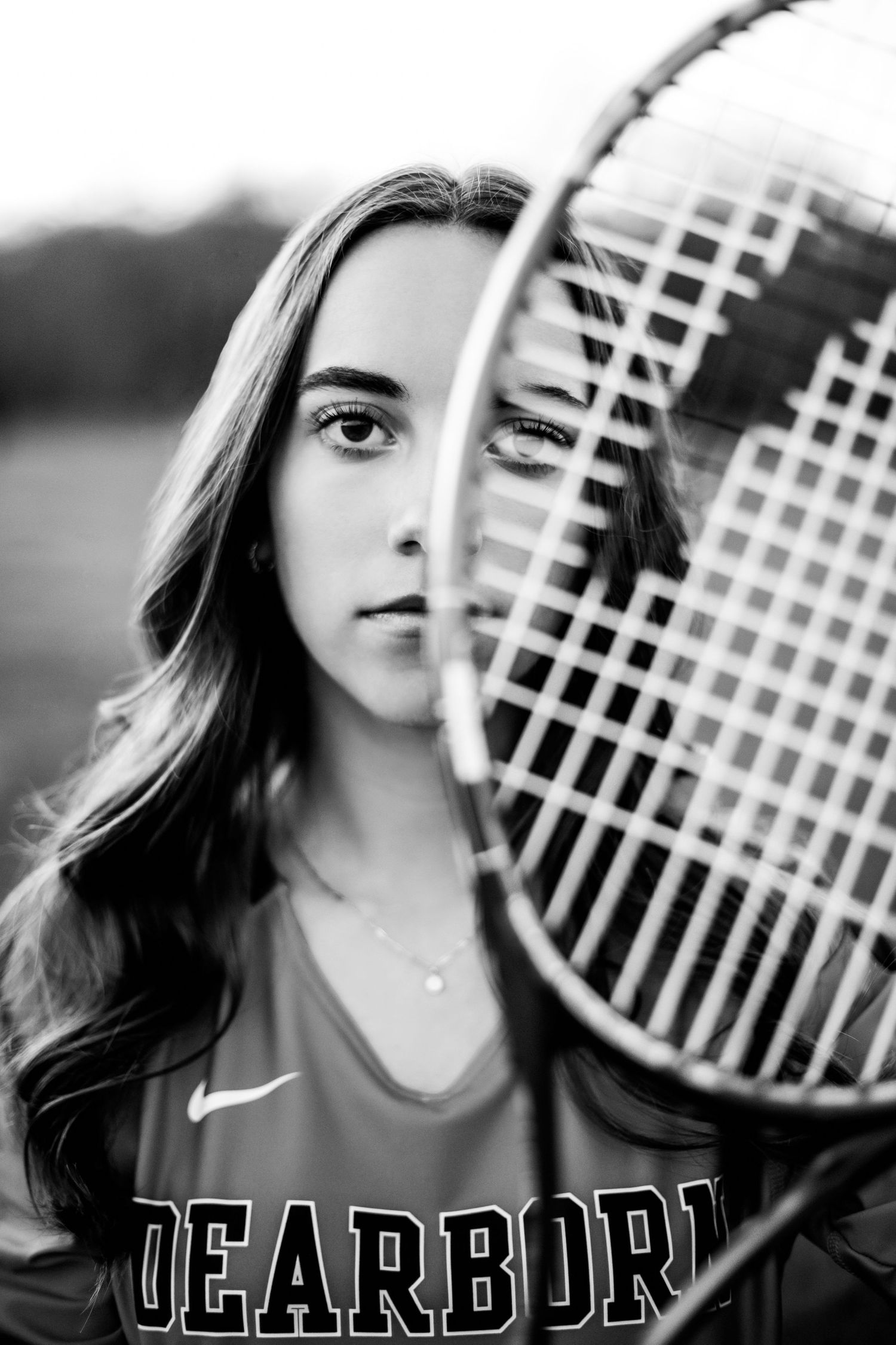 Close-up black and white portrait of a Dearborn athlete with a tennis racket covering half her face.