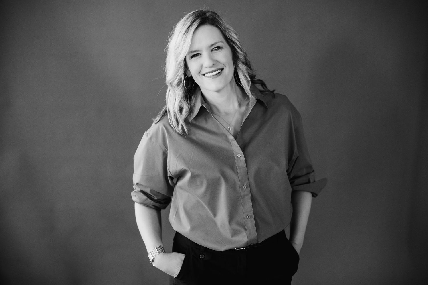 Smiling woman in a button-up shirt poses confidently with hands in pockets in a black and white studio photo.