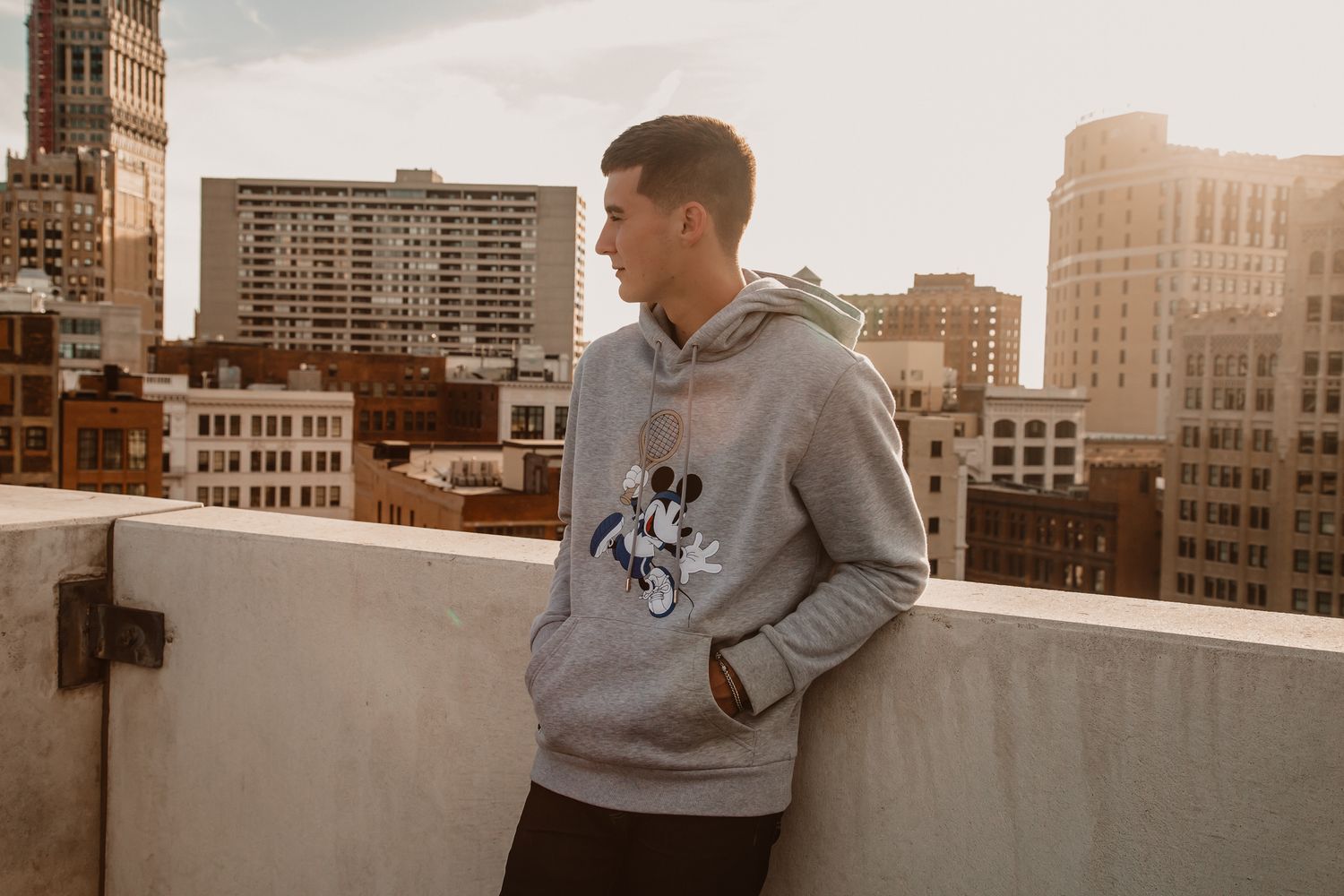 Young man wearing a gray Mickey Mouse hoodie standing on a rooftop with city skyline at sunset.