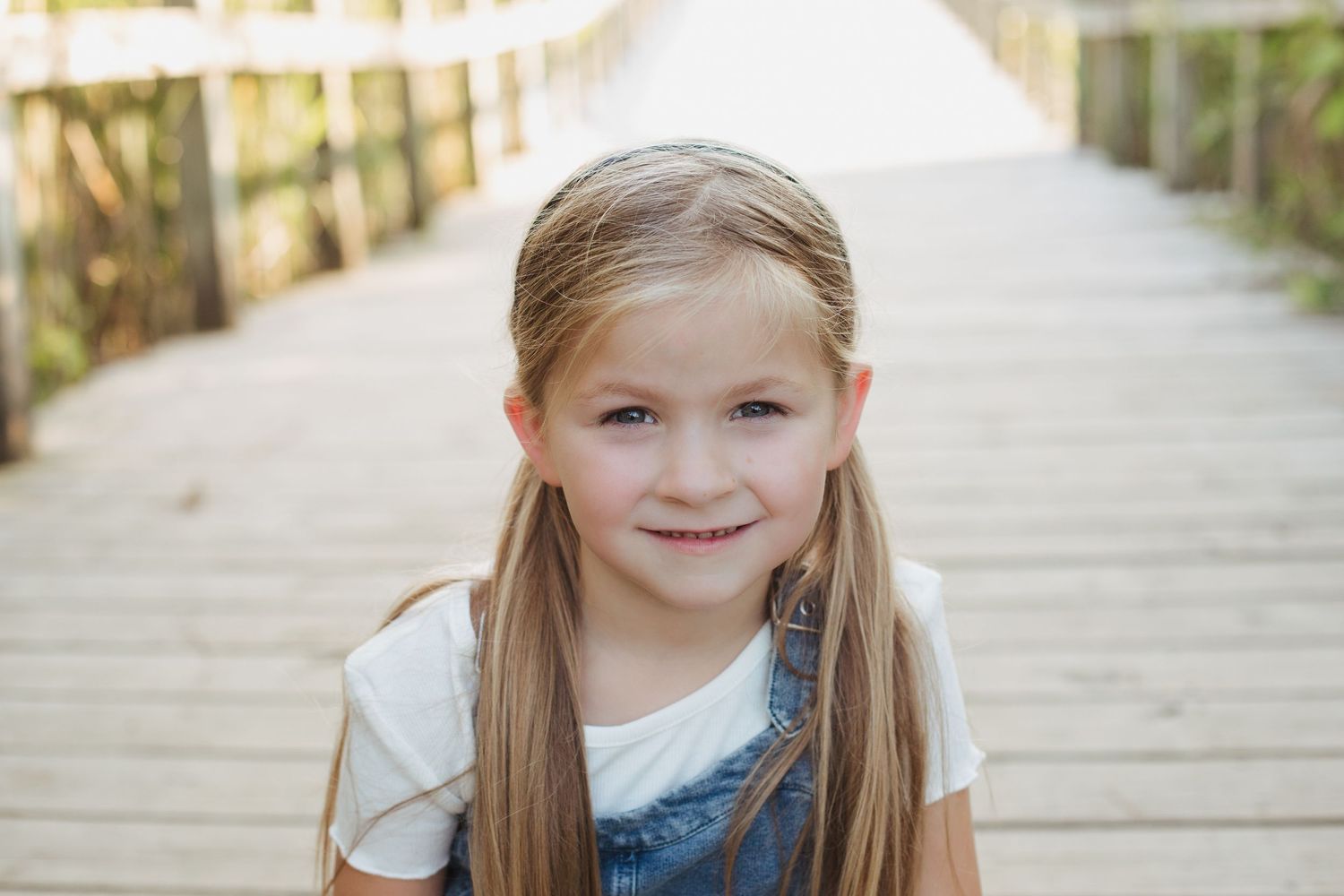 Adorable blonde girl in denim overalls smiles on wooden boardwalk steps outdoors.