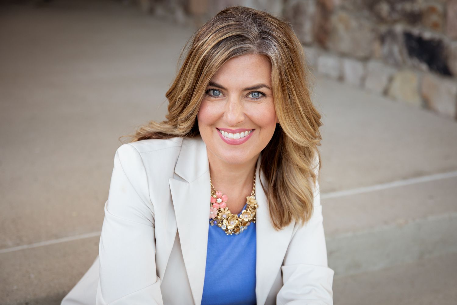 Professional woman smiling in white blazer and blue top with statement necklace, outdoor portrait photo.