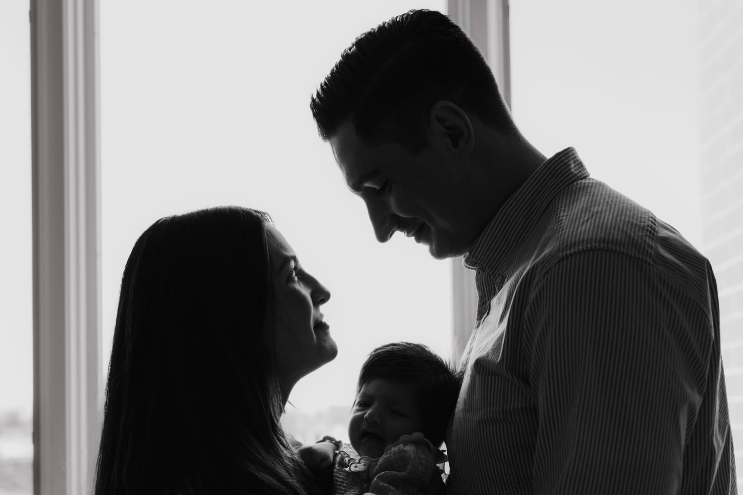Black and white photo of parents holding newborn baby by window light.