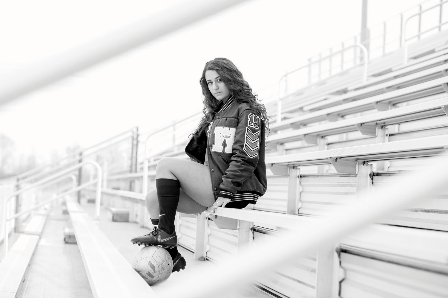 Black and white photo of a young woman in a varsity jacket sitting on bleachers at a sports stadium.