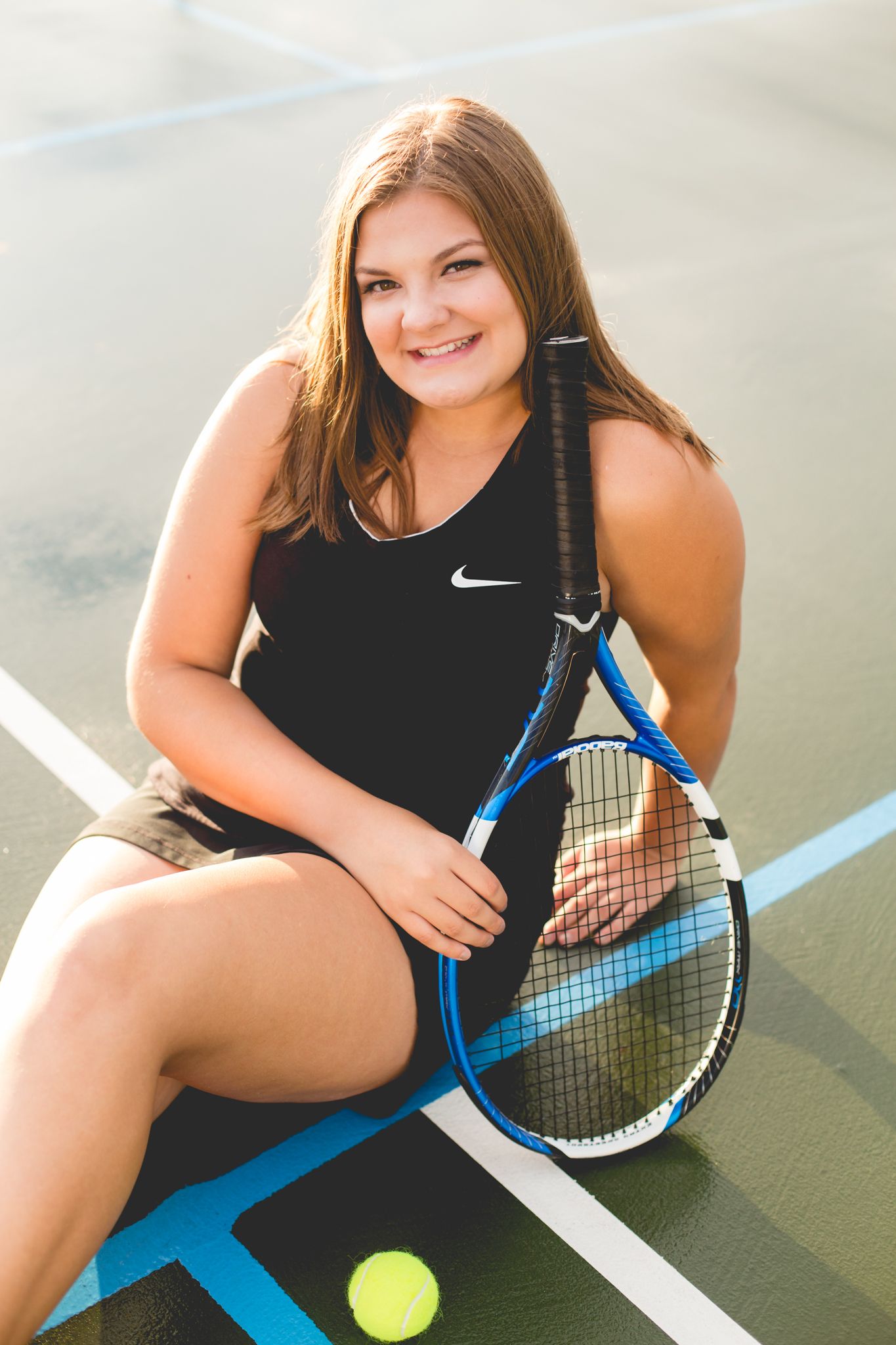 Smiling young woman in black Nike tennis outfit sitting on court holding blue racket with yellow ball nearby.