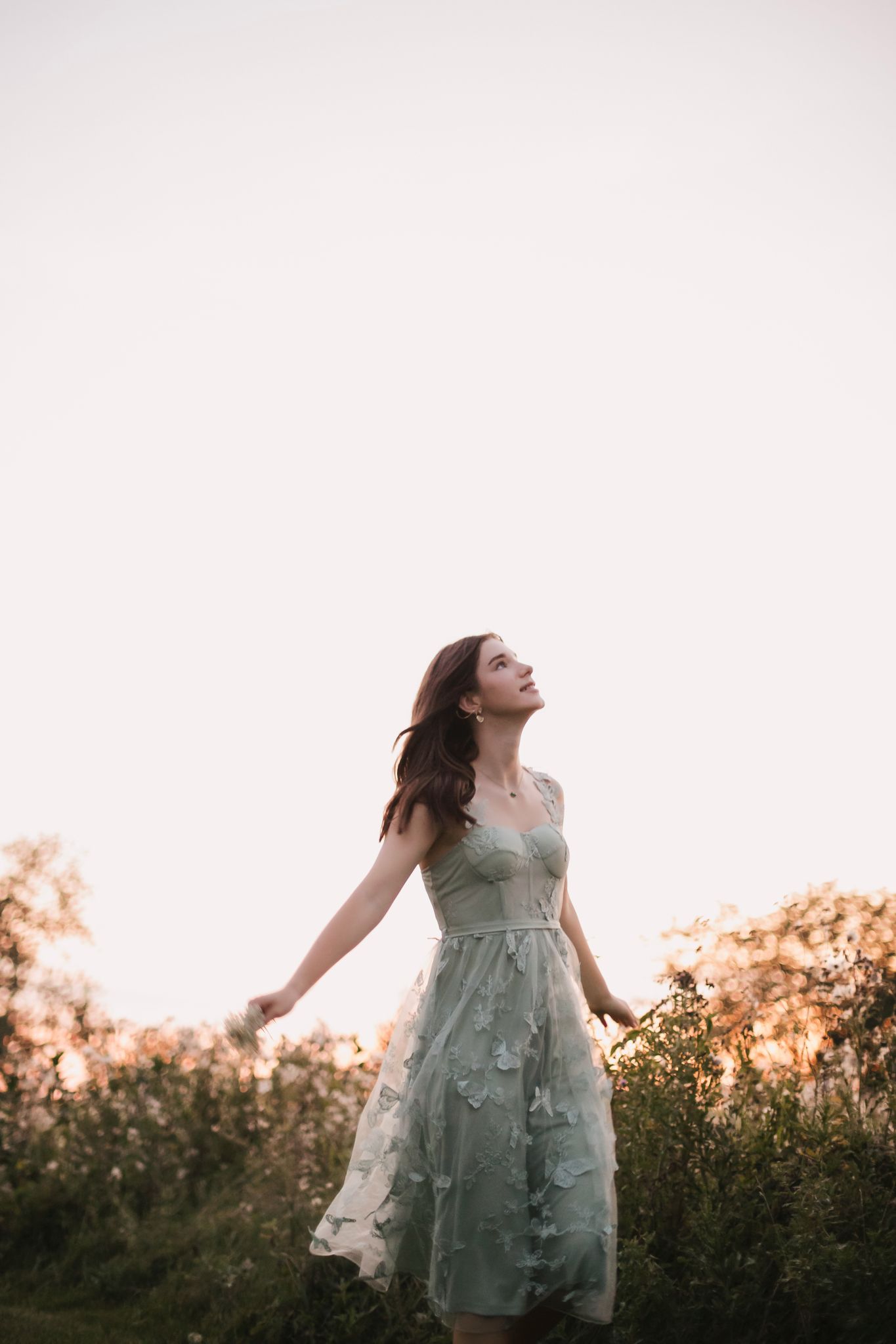 Young woman in sage green floral dress stands in field with arms outstretched at sunset.
