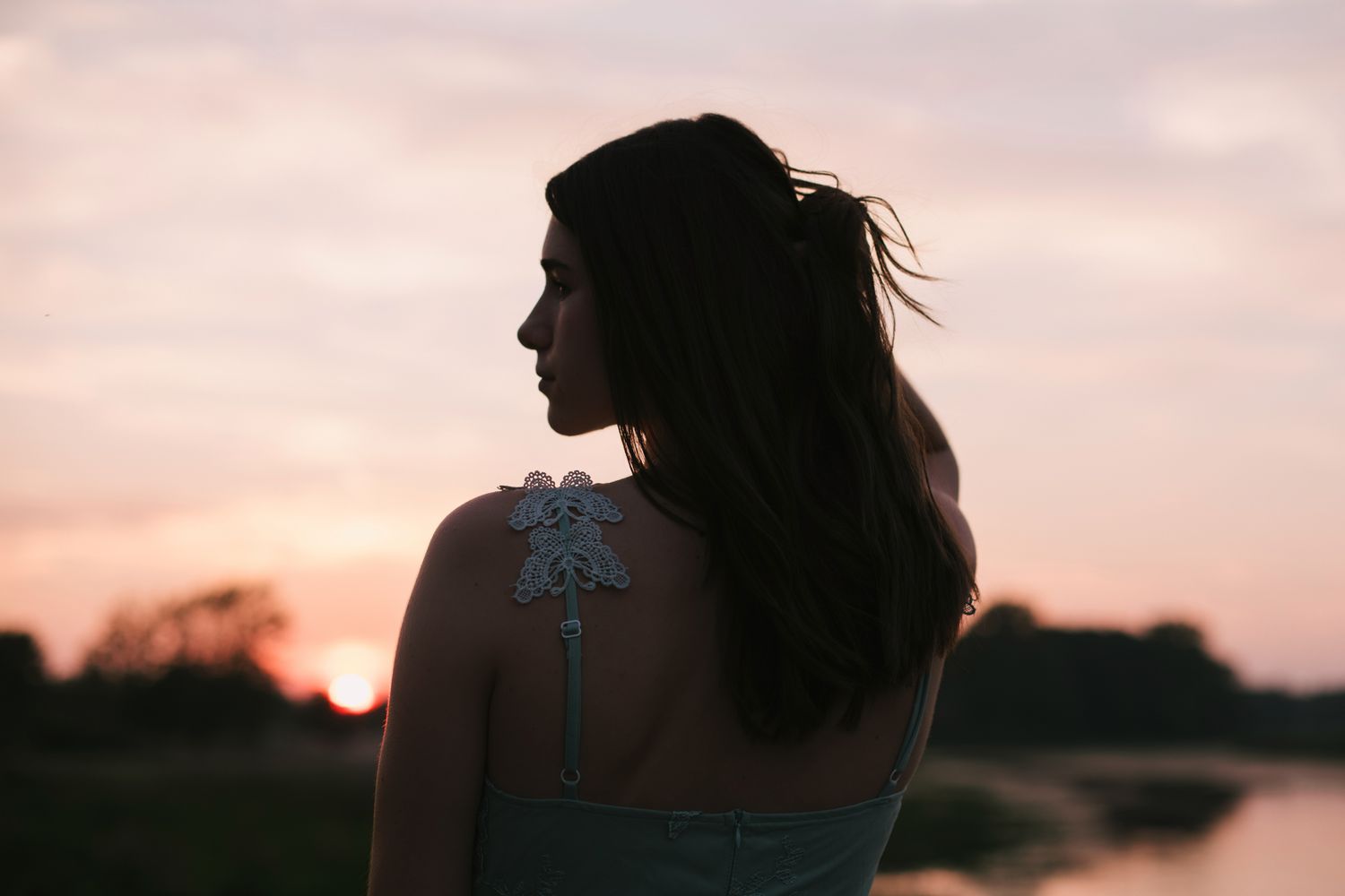 Silhouette of brunette woman in lace back dress at sunset by water.