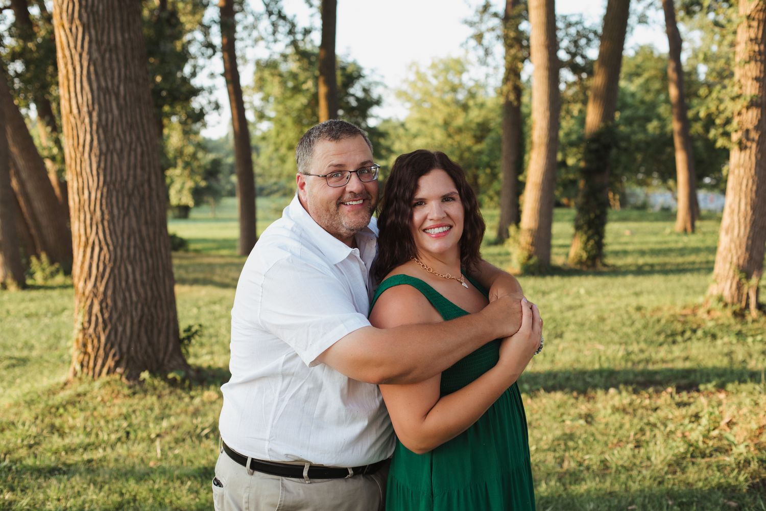 Couple embracing and smiling outdoors in a park, woman in green dress and man in white shirt, surrounded by trees.