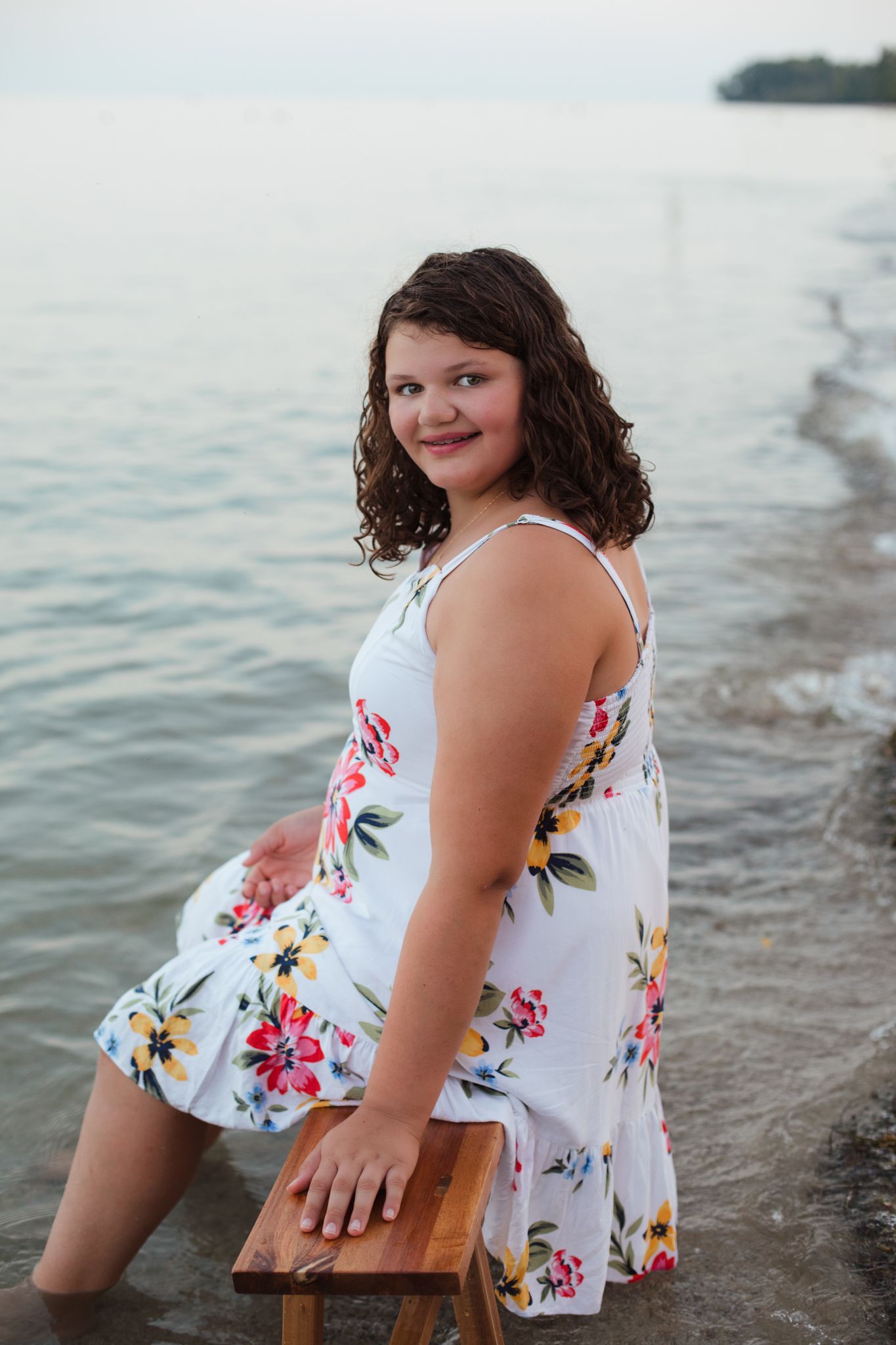 Young woman in white floral sundress smiling by the water's edge, seated on a wooden stool at a lakeside beach.