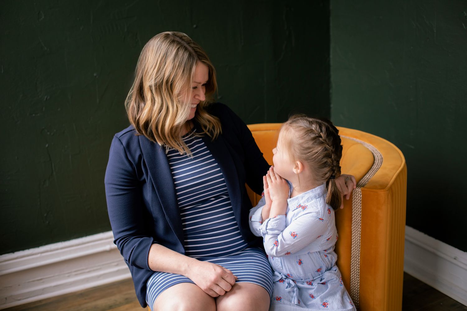 Pregnant woman in a striped dress looks lovingly at a young girl sitting beside her in a yellow chair.