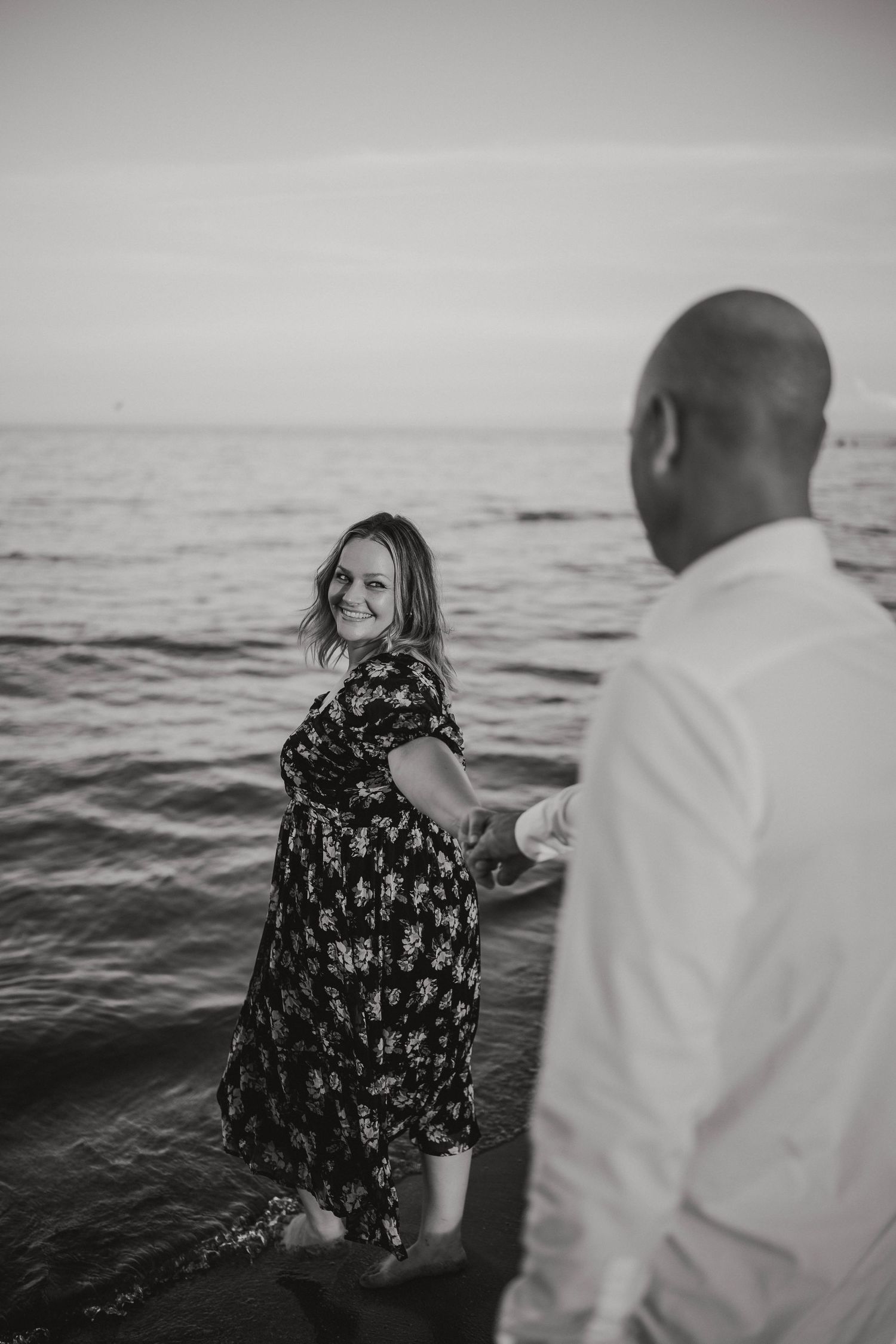 Black and white beach photo of woman leading man by hand into the ocean.