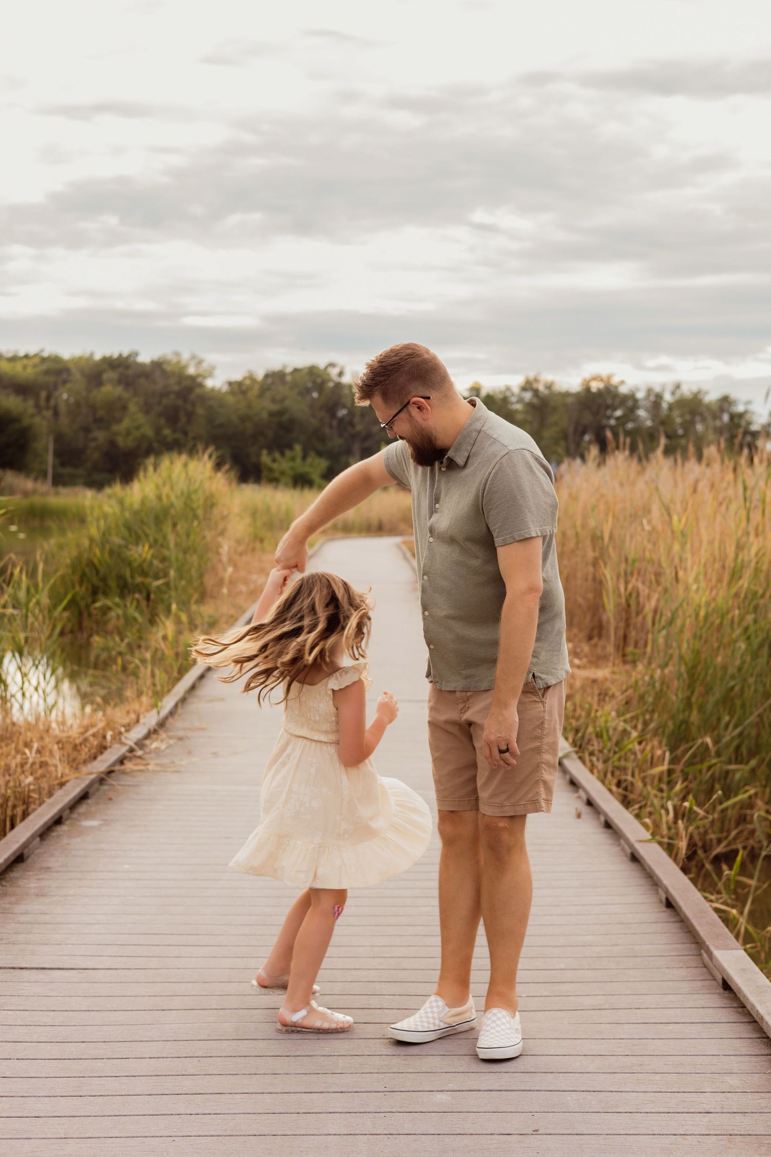 A father twirls his young daughter in a white dress on a wooden boardwalk surrounded by tall grass and trees.