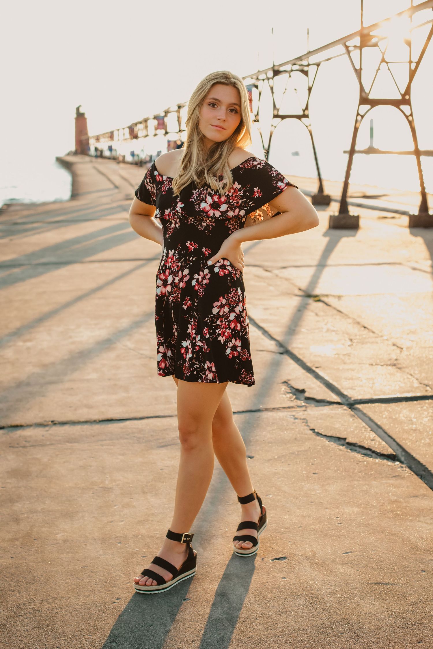 Young woman in black floral mini dress and wedge sandals posing on a sunlit pier.