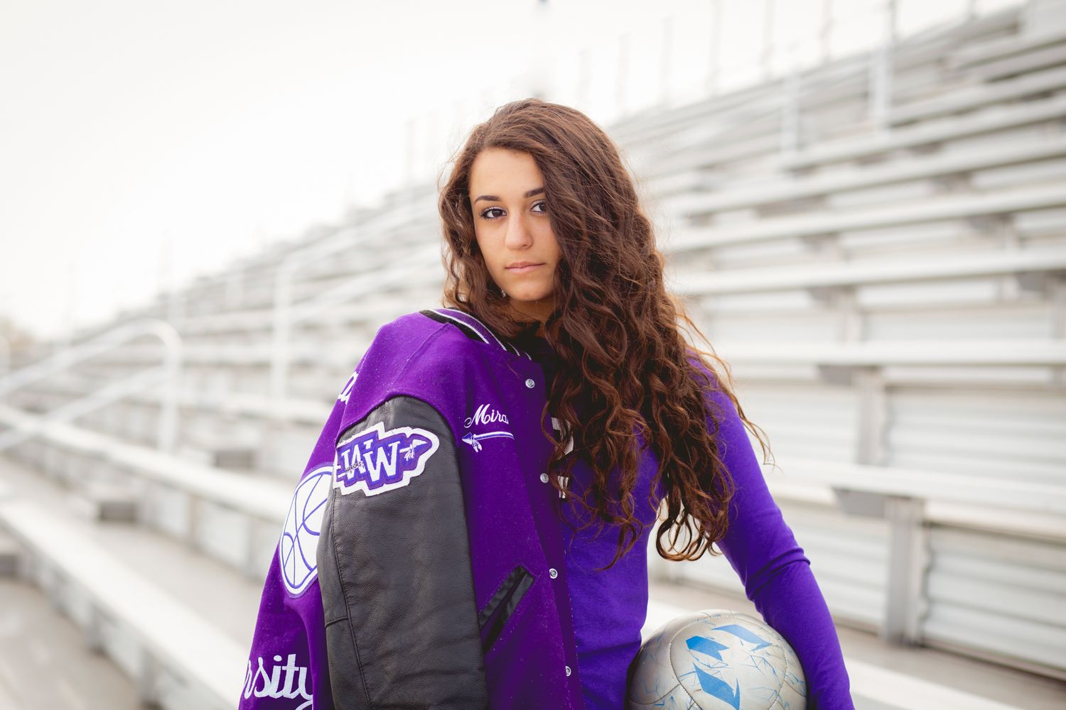 Curly-haired teenage girl holds soccer ball wearing purple varsity jacket on bleachers for senior portraits.