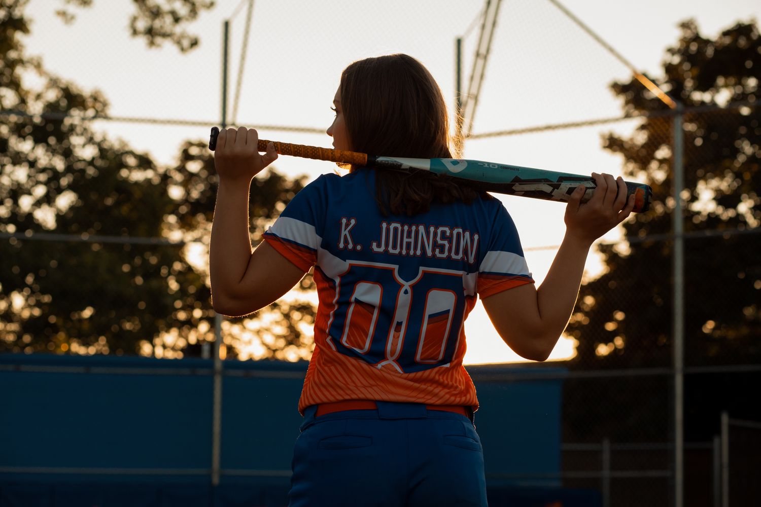 Softball player wearing K. Johnson number 00 jersey holding a bat across her shoulders at sunset.