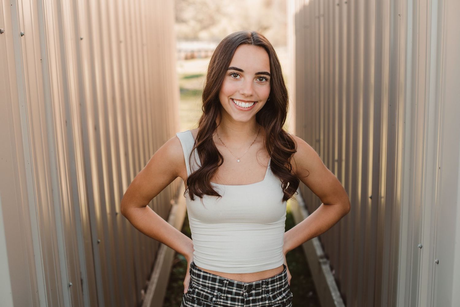 Smiling brunette teen in white crop top and plaid shorts posing between corrugated metal walls outdoors.