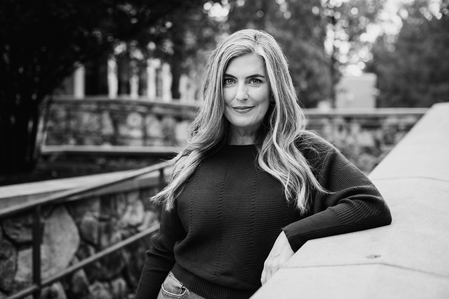 Black and white portrait of a blonde woman in a dark sweater leaning against a wall outdoors.
